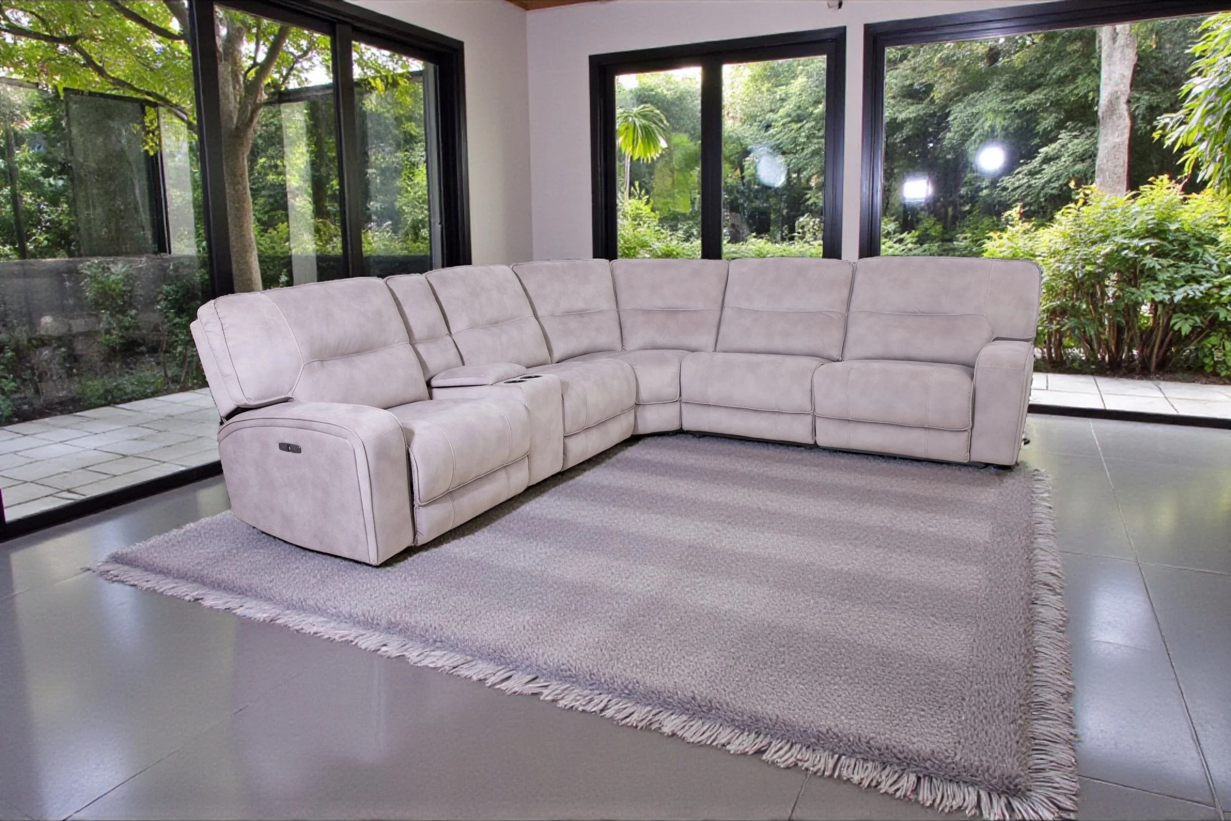 A modern living room with a beige sectional sofa, large arched windows with white curtains, a geometric mountain painting on the wall, a black side table with a small lamp and plant, and a patterned area rug on a dark wood floor.