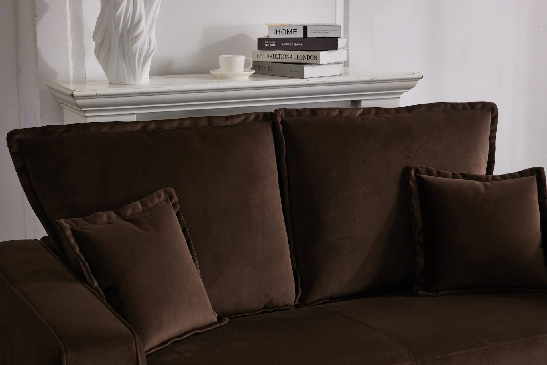 A dark brown velvet sofa with matching cushions in front of a white fireplace mantel, which has a decorative white vase, a stack of books, and a ceramic teacup and saucer on top.