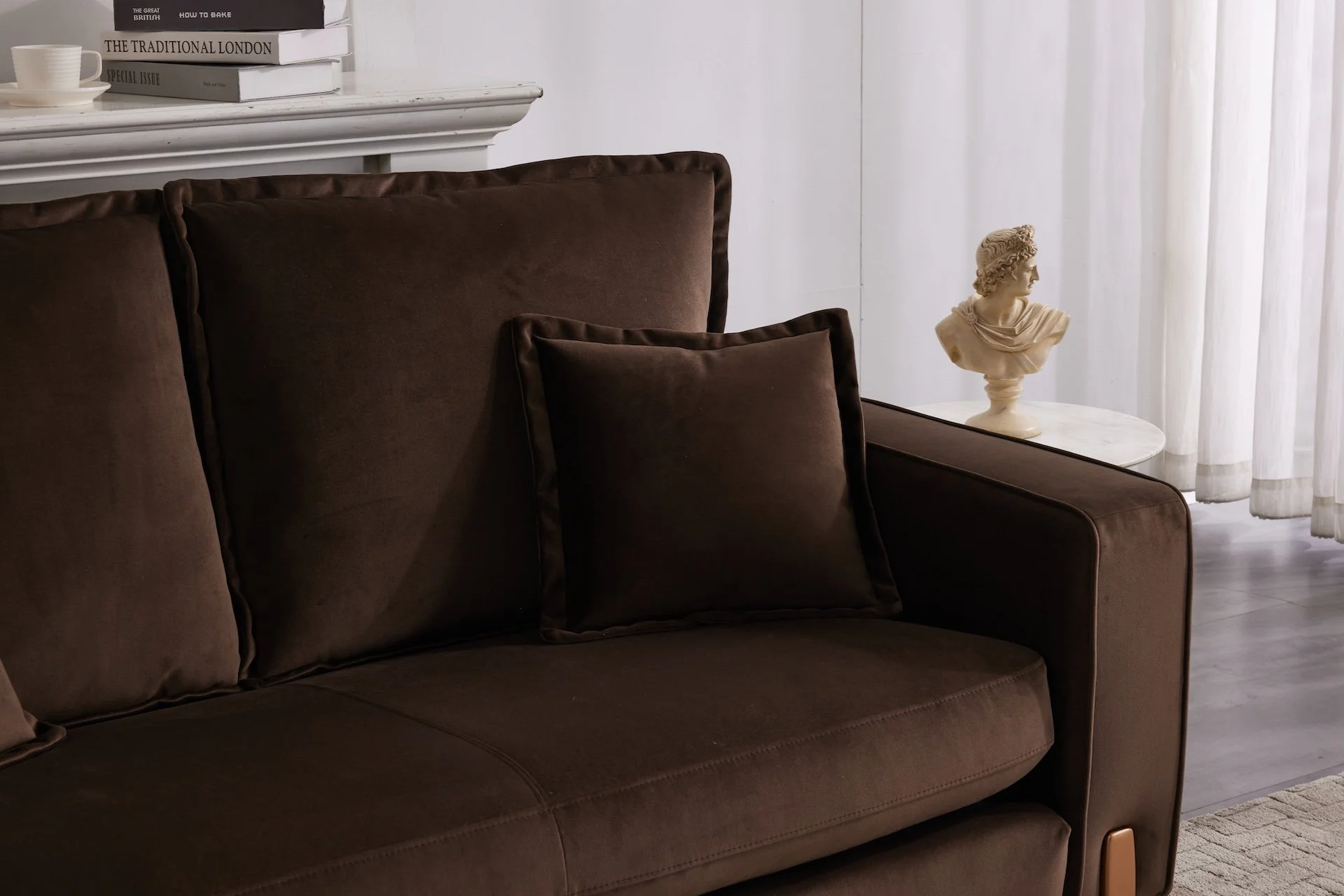 Close-up of a brown sofa with matching cushions in a living room, with a white mantel above, a stack of books, and a small marble-topped side table with a bust sculpture near a window with curtains.
