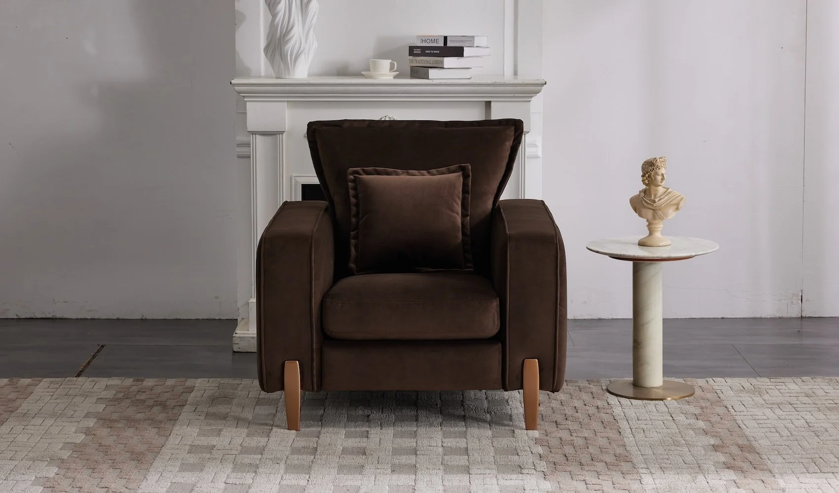 A cozy living room corner with a dark brown upholstered armchair with a matching pillow, a small round marble-topped side table holding a classical bust sculpture, a white fireplace mantel decorated with books, a white vase, and a cup, on a beige and