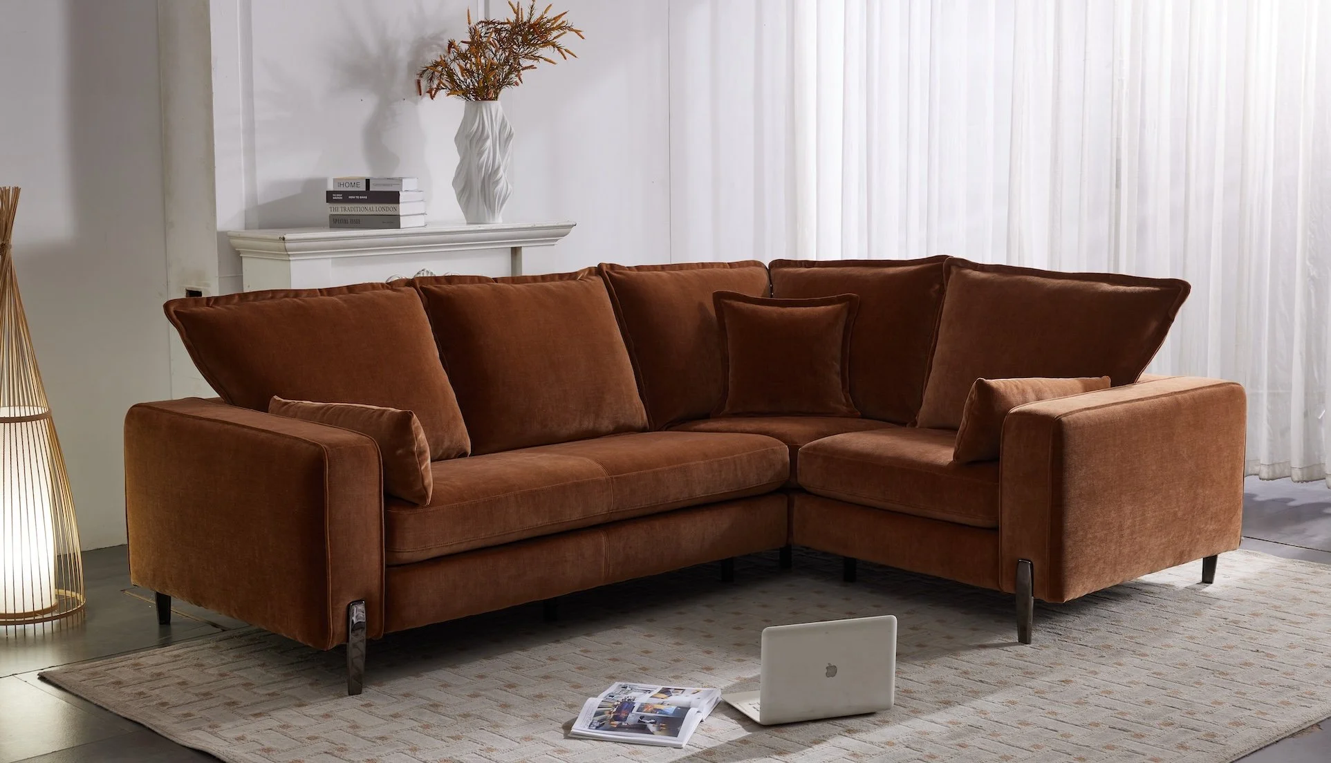 A brown L-shaped sofa with cushions in a living room with white curtains, a beige rug, a white decorative shelf with books and a vase with dried flowers, and a laptop on the rug.
