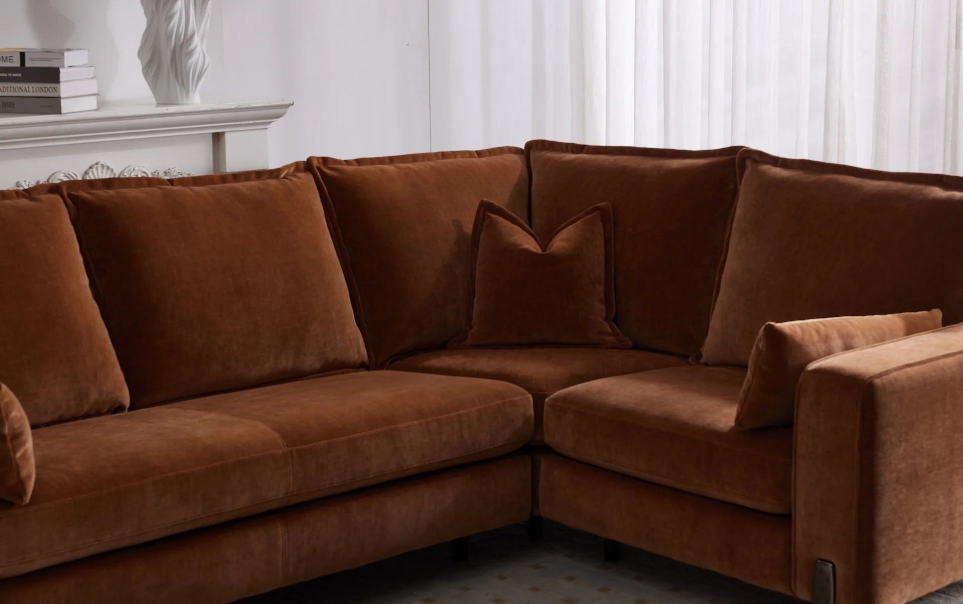 A brown sectional sofa with rectangular and throw pillows, located in front of a window with sheer white curtains, in a living room with white walls and a decorative white fireplace mantel with books and a vase.
