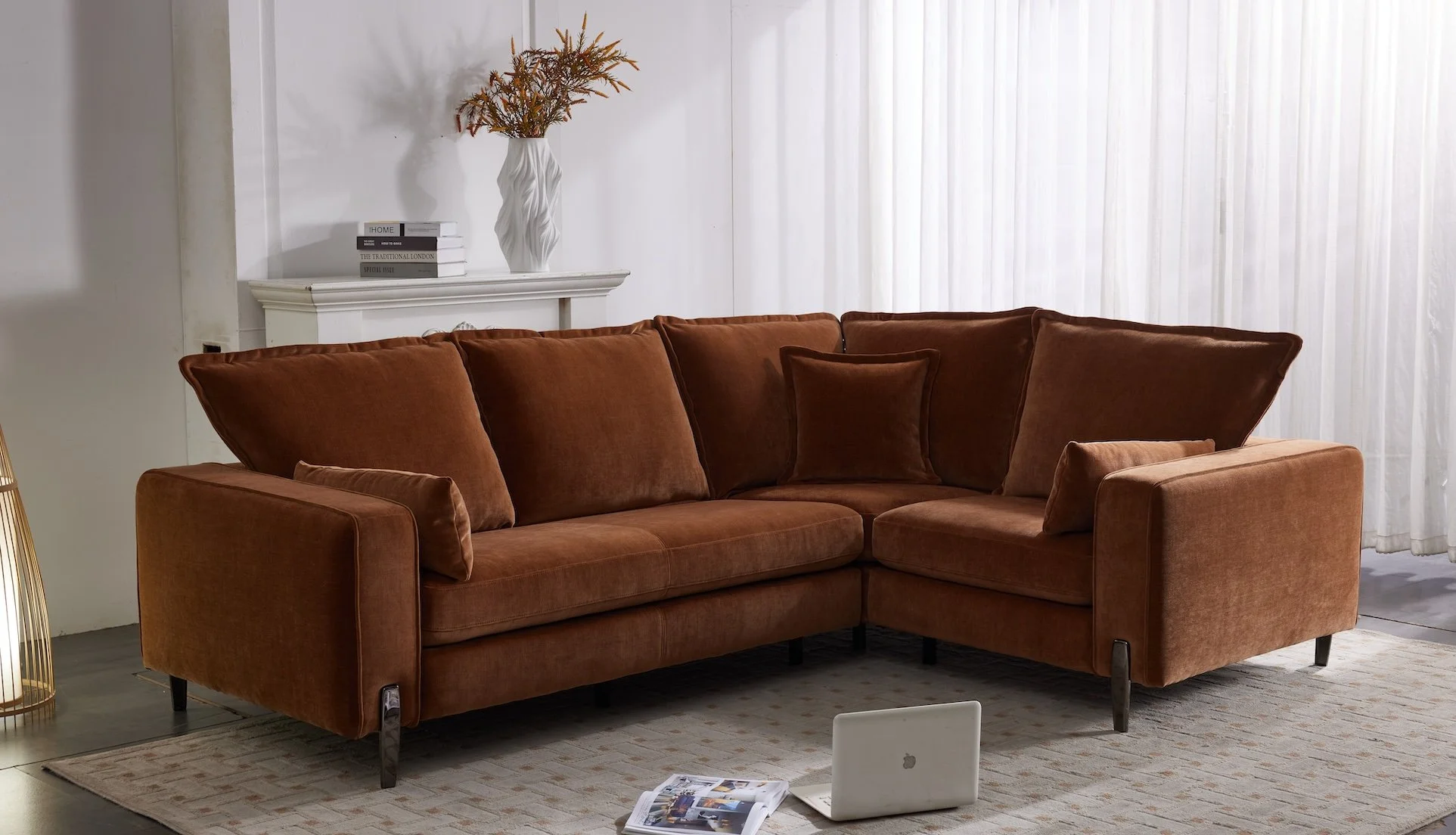 Living room with a rust-colored sectional sofa, a white laptop, along with a magazine, on a rug. Behind the sofa, white curtains cover a window, and a white fireplace mantel holds a tall white vase with dried flowers and some books.
