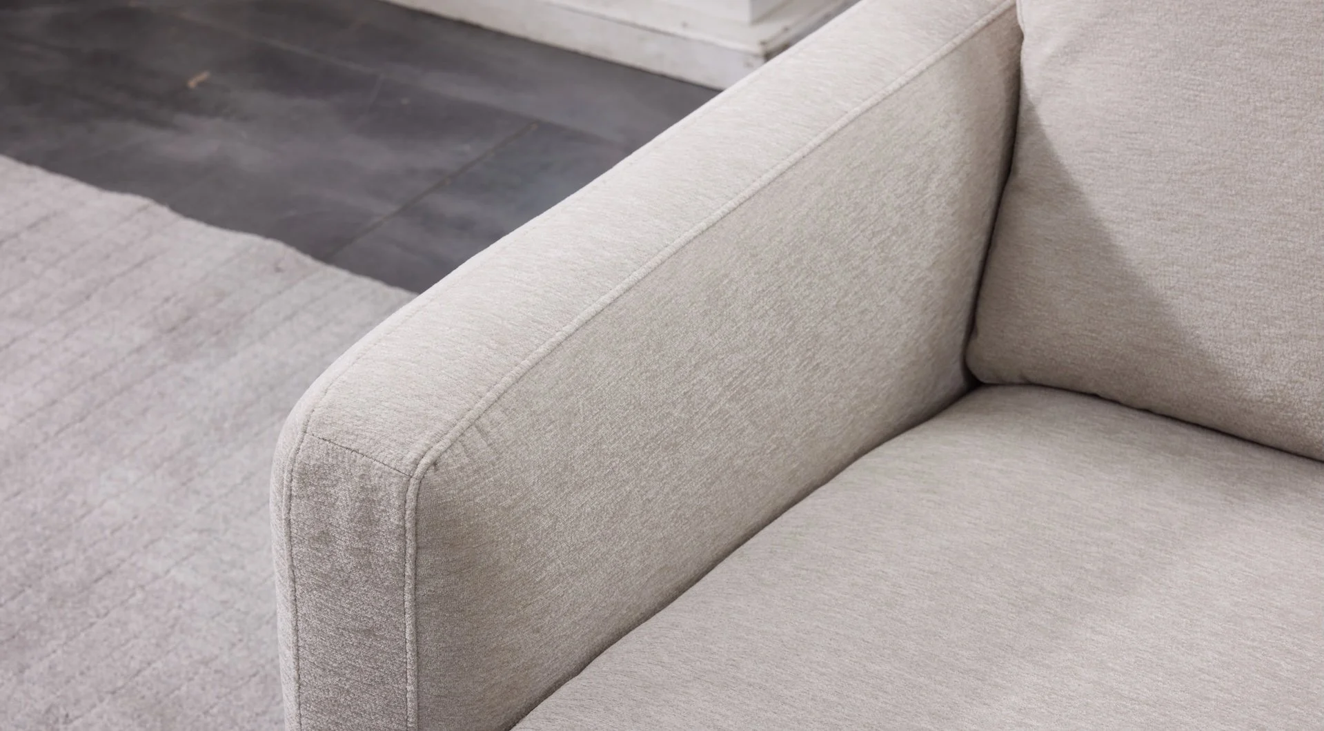 Close-up of a beige upholstered sofa armrest and cushion with a dark wood and tile floor in the background.