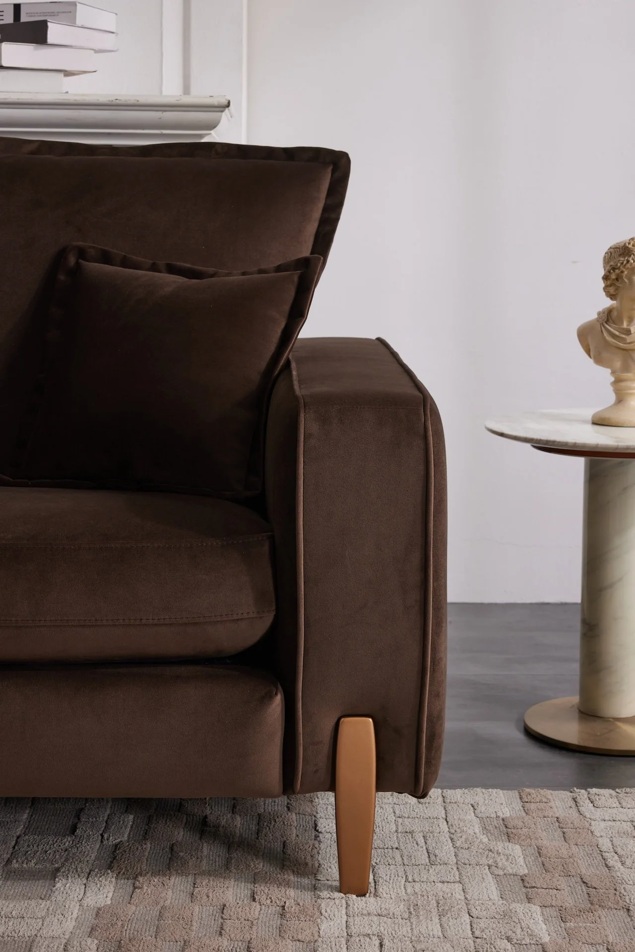 Close-up of a brown velvet sofa with two matching pillows and a wooden leg, beside a marble side table with a classical bust on top, on a textured beige rug and dark wooden floor.