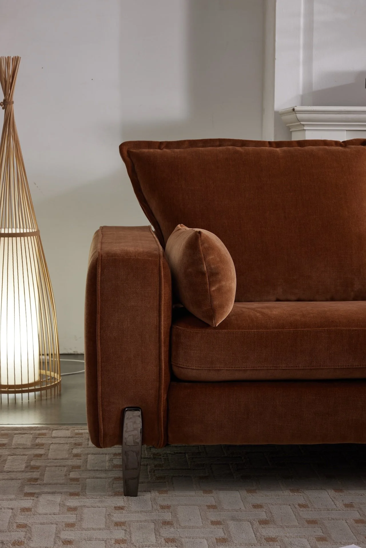 A brown velvet couch with a matching pillow, next to a tall decorative floor lamp with a warm glow, placed on a beige patterned rug in a cozy living room.