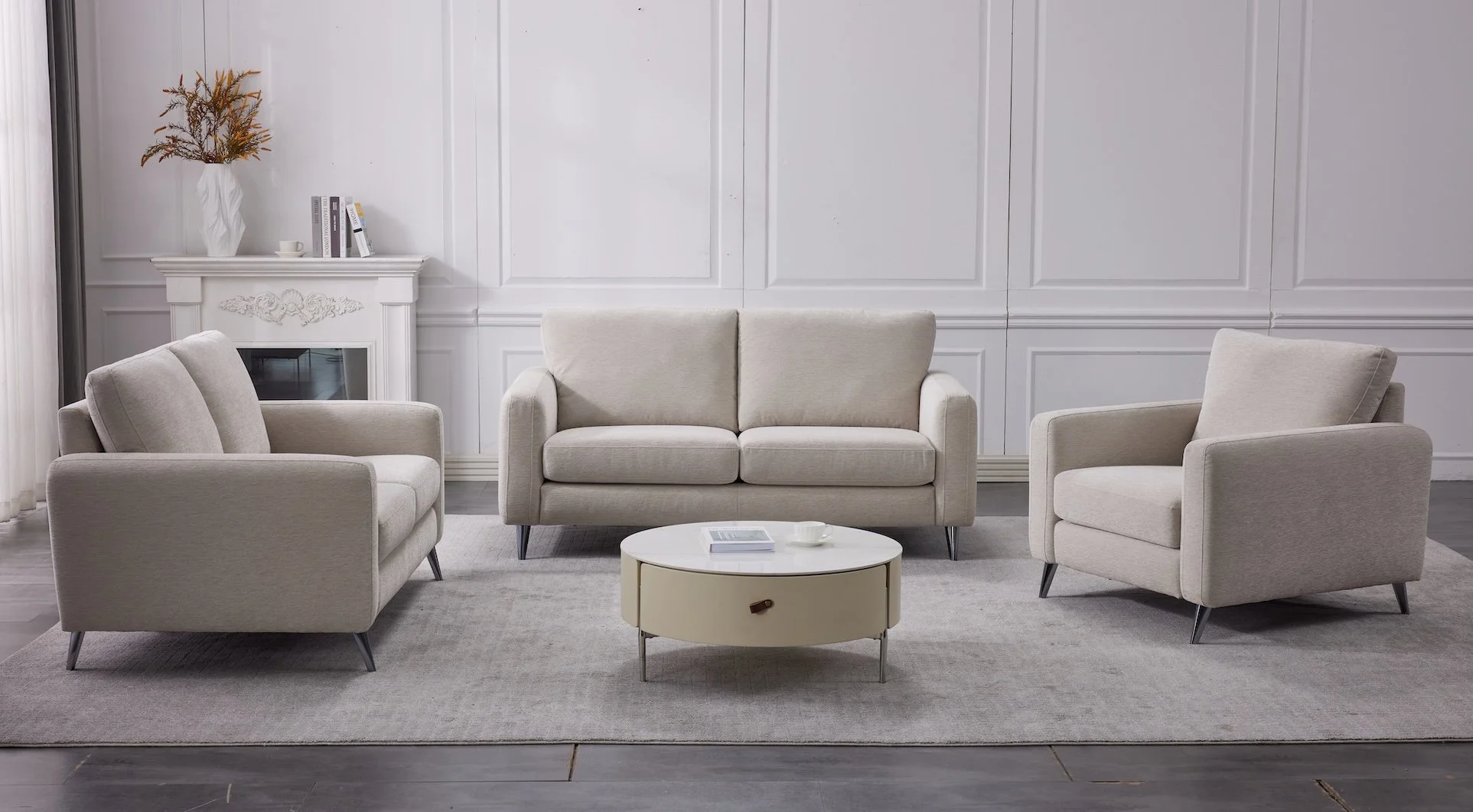 Living room with three beige sofas, a white round coffee table, a gray area rug, white walls with decorative panels, a fireplace with books and a vase on top, and a curtain on the left side.