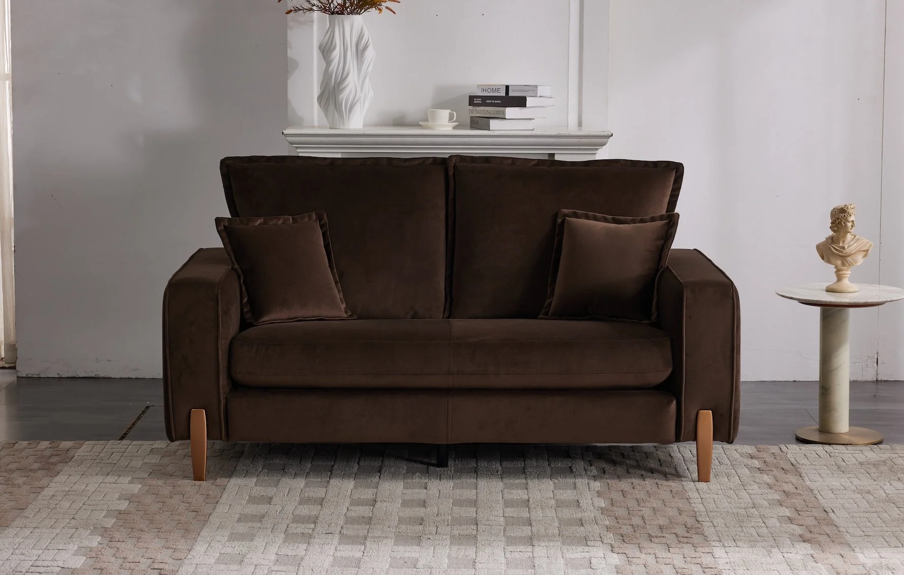 A brown suede sofa with four matching pillows, positioned on a patterned area rug in front of a white wall. On the wall above the sofa, there is a white mantel with decorative items, including a white ceramic vase with floral design, a small stack of