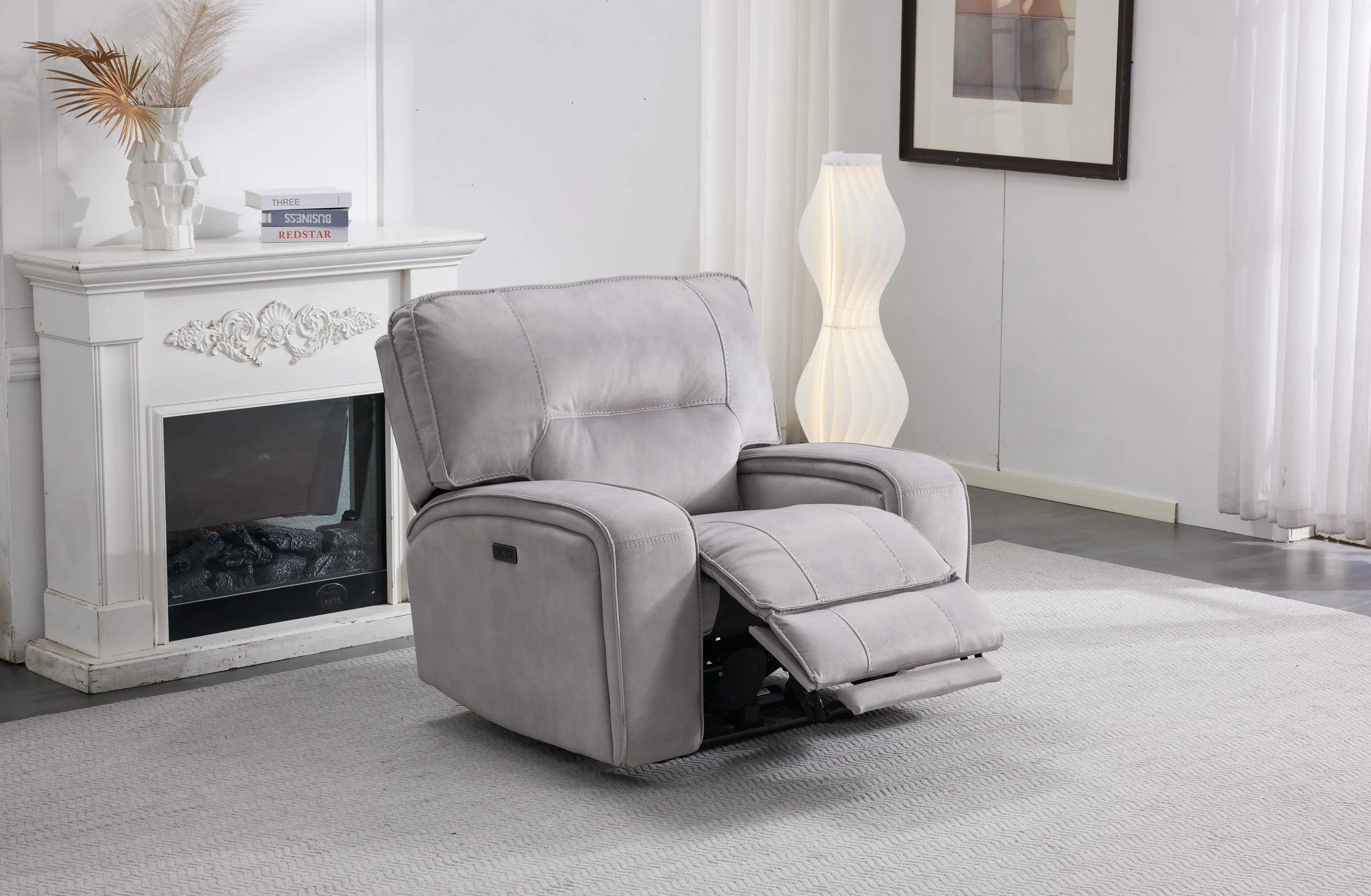 A cozy living room corner with a gray recliner chair, a white fireplace with decorative molding, a white tall floor lamp with a wavy design, and a white area rug. There are also plants and books on the mantel, and a framed picture on the wall.