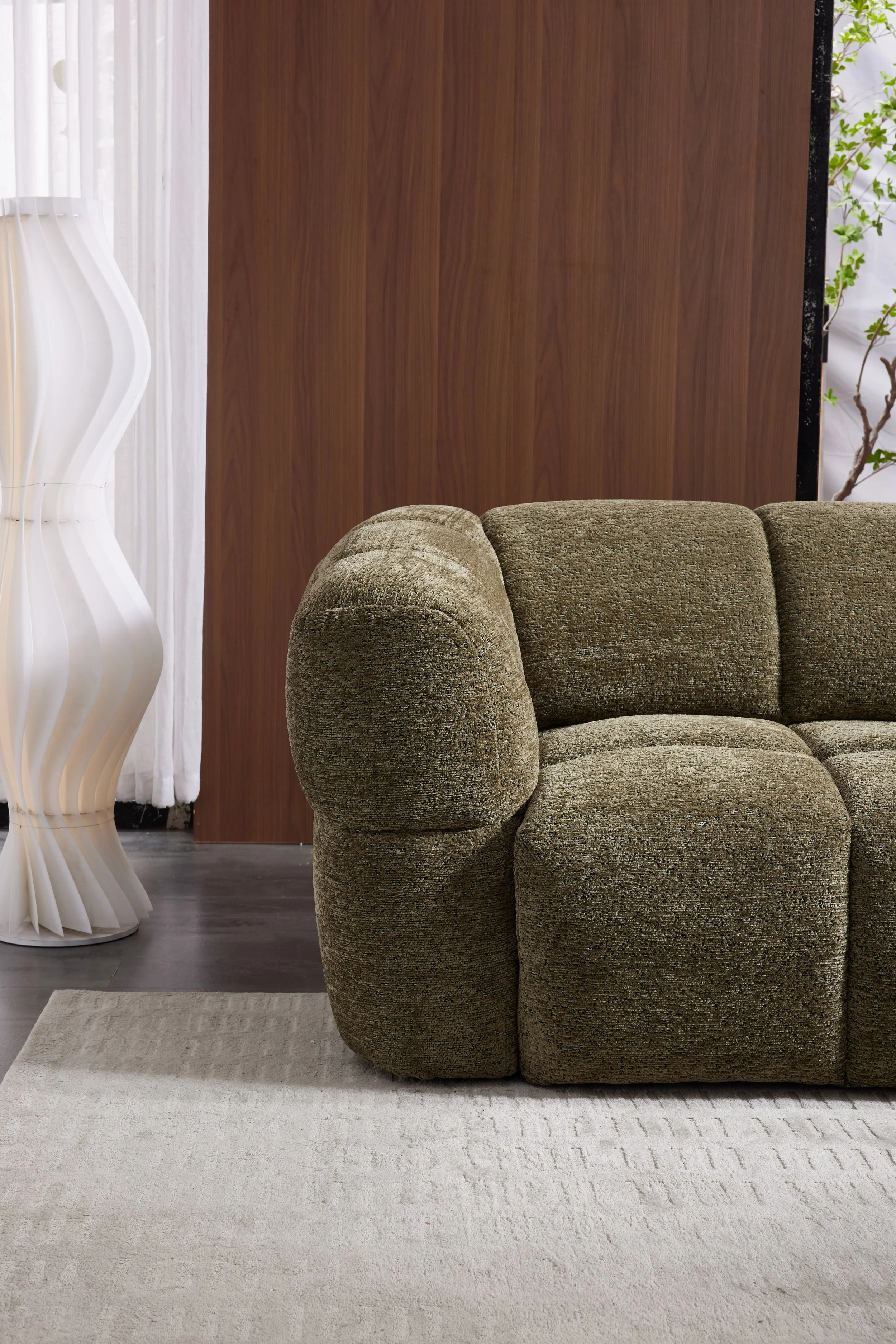 A living room scene with a textured olive-green armchair, a beige area rug, a wooden partition, a white sculptural floor lamp, a sheer white curtain, and some green plants in the background.