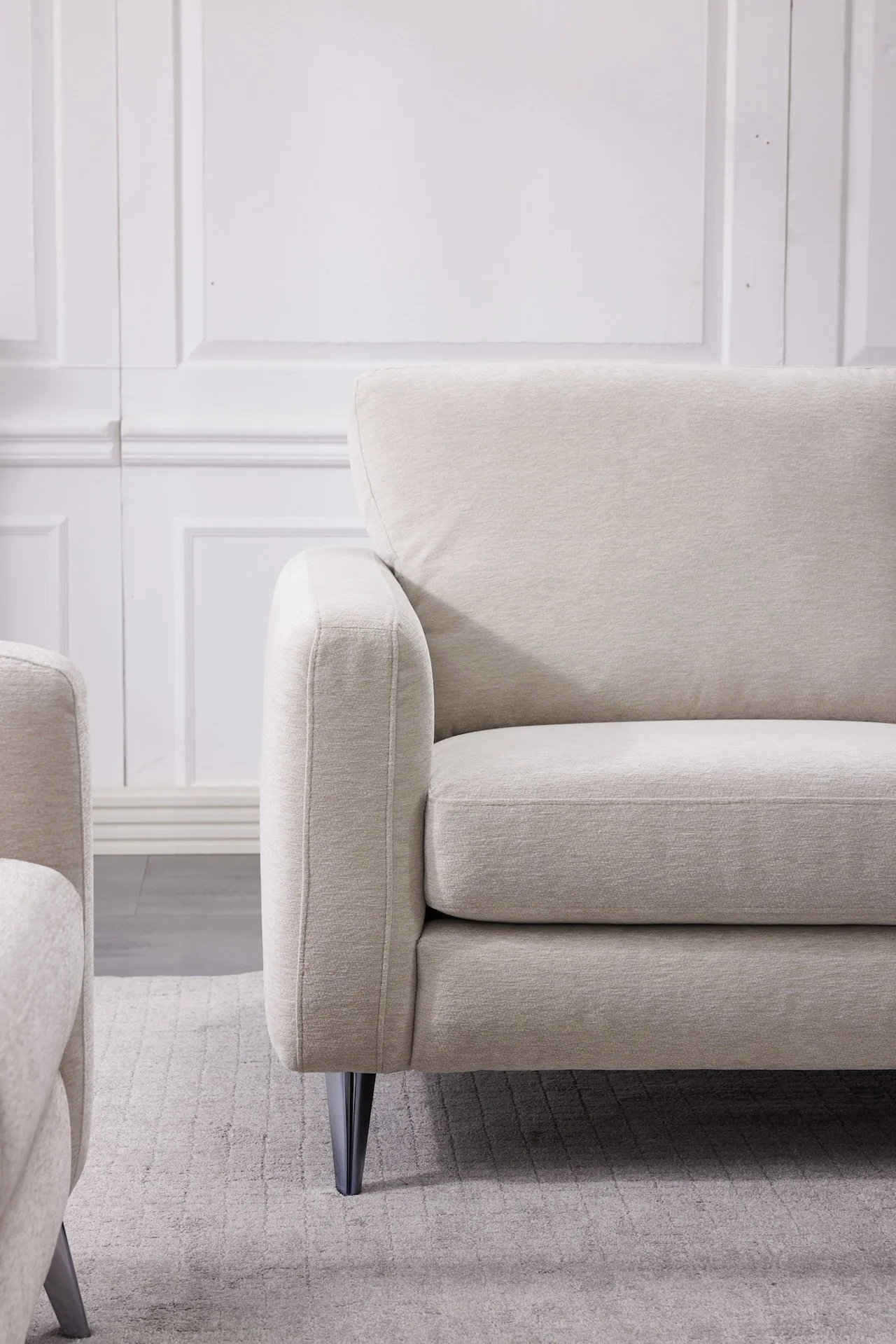Part of a beige sofa with a dark metal leg, placed on a beige textured carpet in a room with white walls and wainscoting.