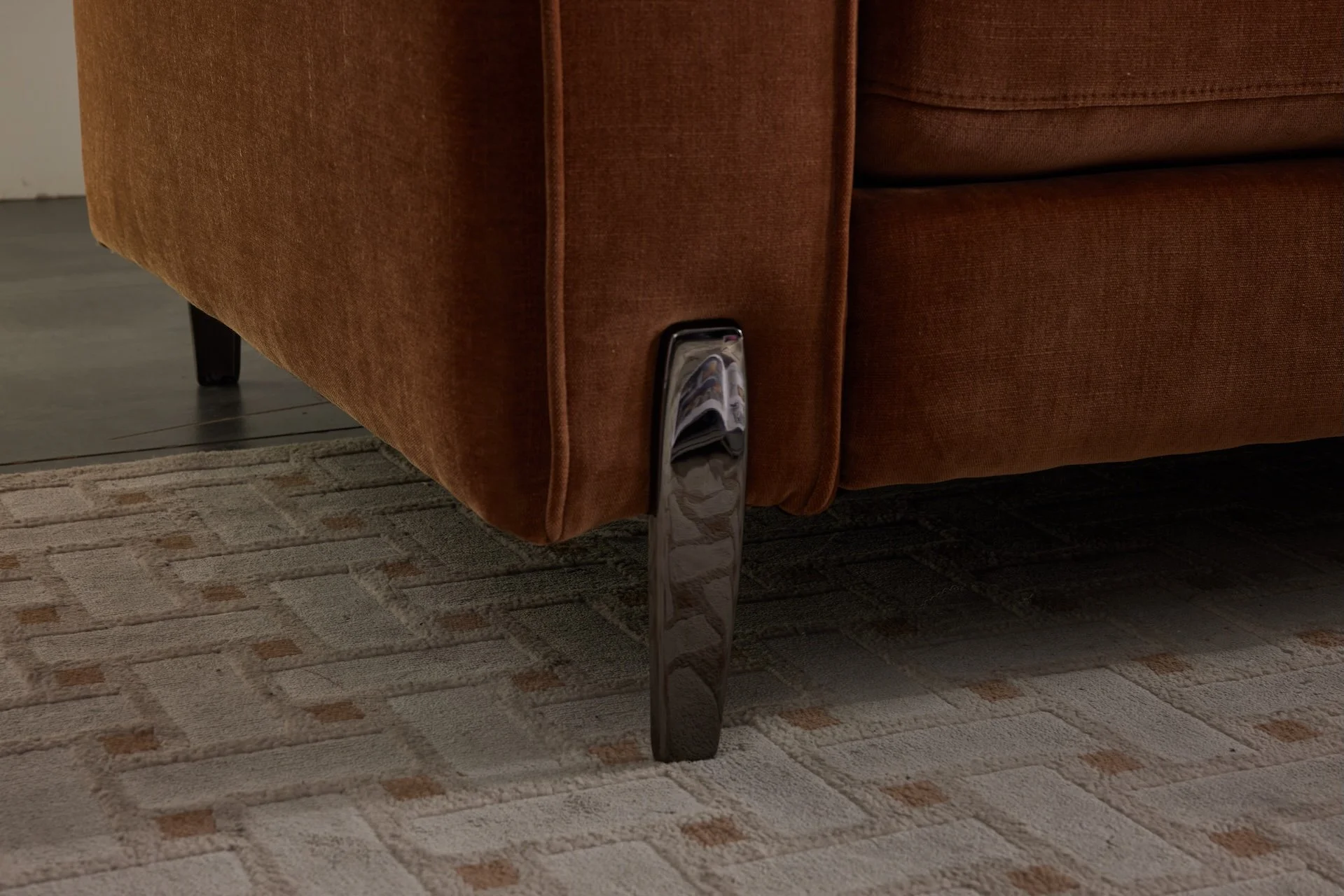 Close-up of a brown upholstered sofa with a shiny black metal leg on a patterned beige and brown area rug.