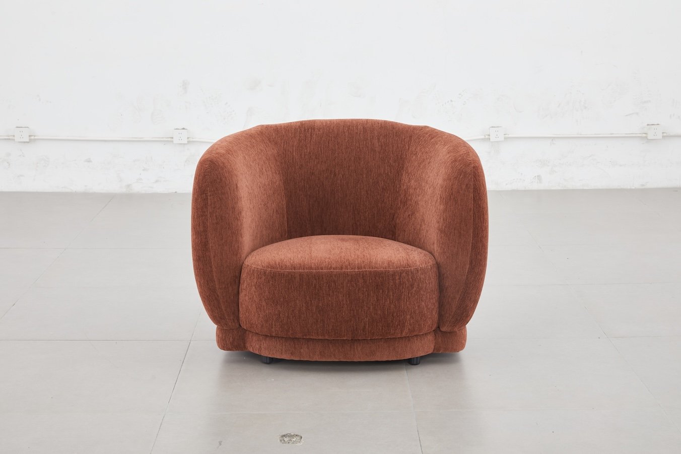 A single rounded rust-colored upholstered armchair in a minimalist space with white walls and a light gray floor.