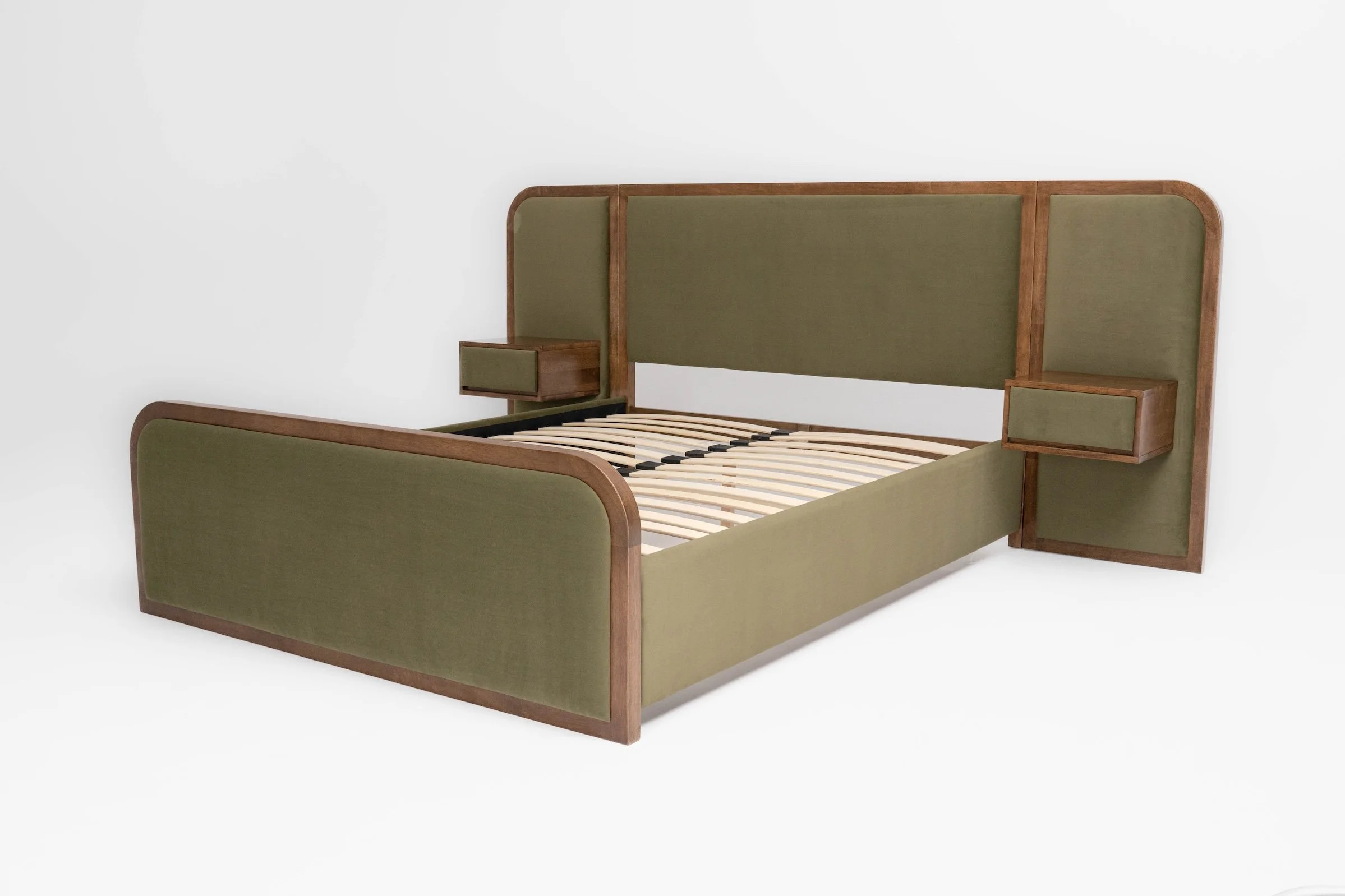 A bedroom with a wooden bed frame, green upholstery, white bedding, and four pillows. There's a small wall shelf with a vase of purple flowers, a framed line drawing, and a window with yellow curtains.