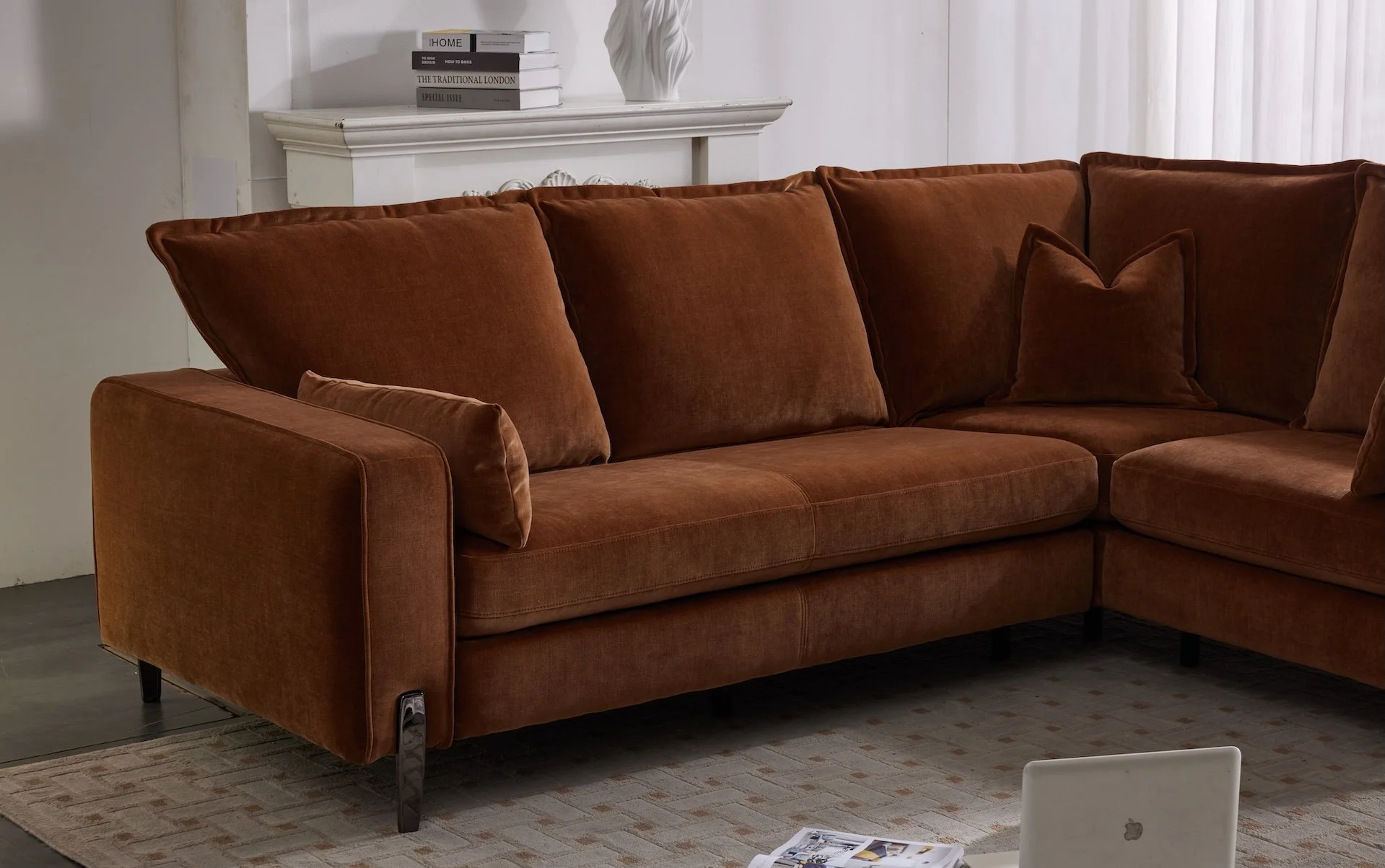 A brown sectional sofa with multiple cushions in a living room setting. There is a stack of books and a decorative vase on a white fireplace mantle in the background, and a portion of a laptop and magazines on the carpet in the foreground.