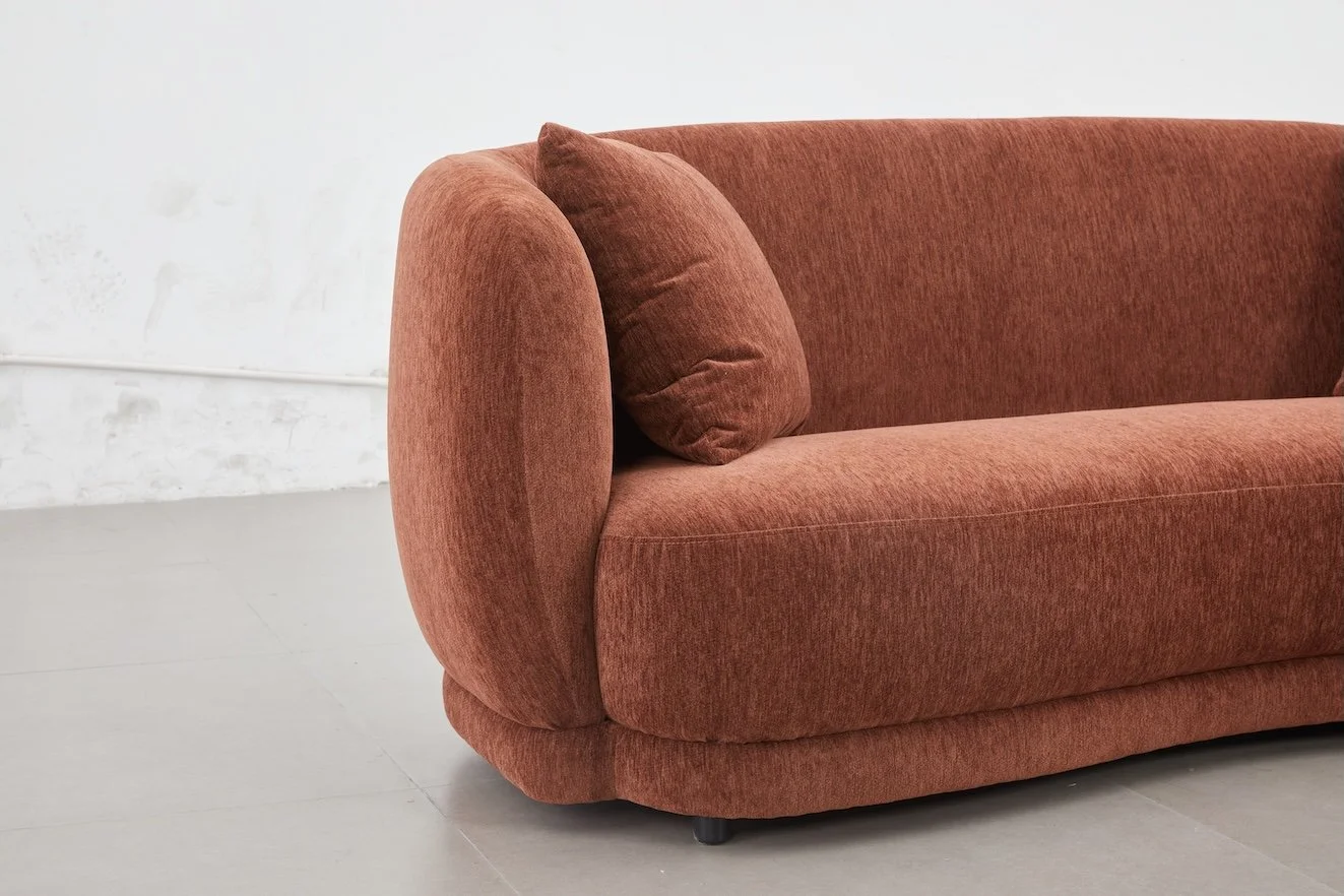 A rust-colored, plush fabric sofa with a matching cushion on a light-colored floor with a plain white wall in the background.