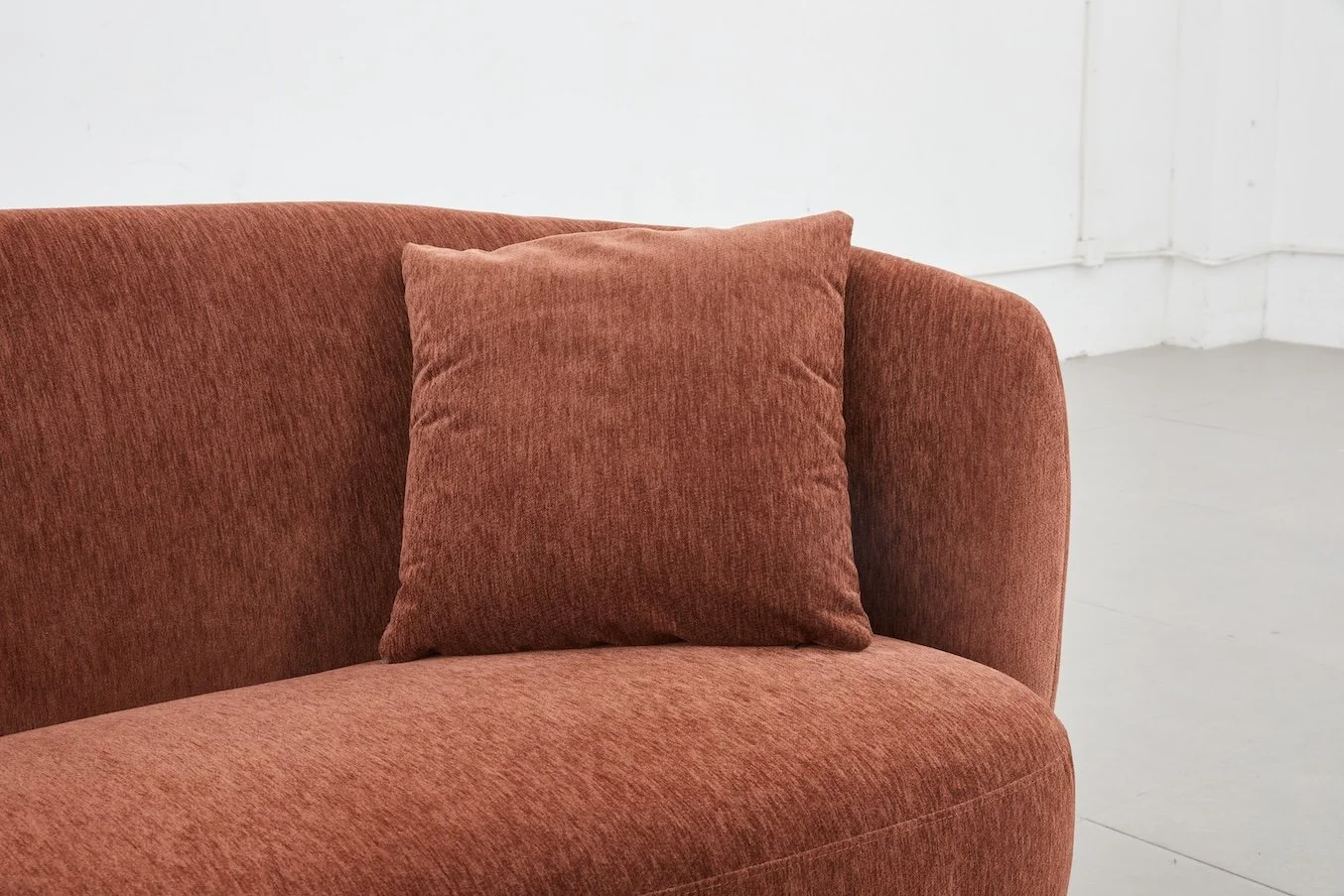 A rust-colored fabric sofa with a matching cushion, set against a white wall and white floor.