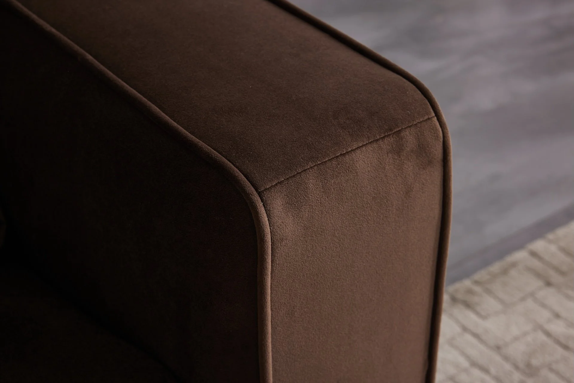 Close-up of a dark brown upholstered armrest of a sofa against a grayish wooden floor.