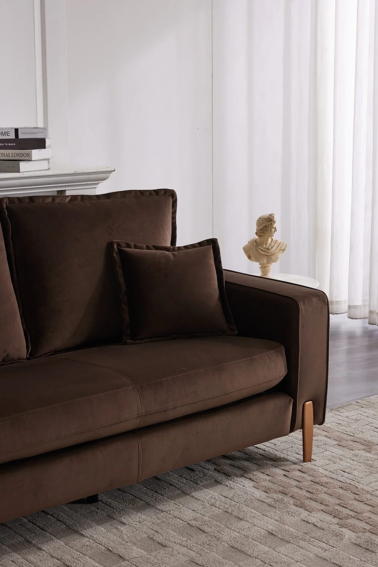 A dark brown sofa with matching throw pillows, a stack of books on a white mantle, a small bust sculpture on a side table, and sheer white curtains in the background.