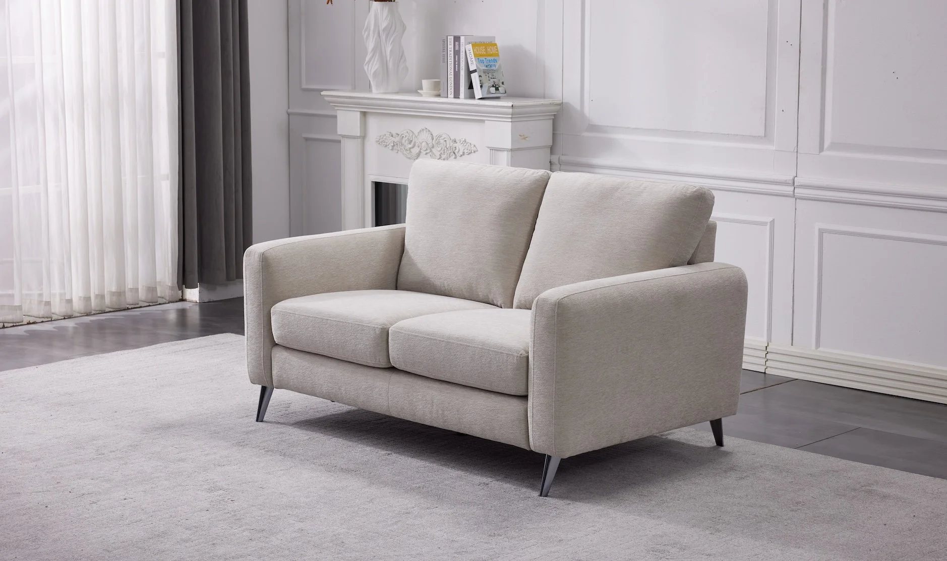 A beige two-seater sofa with angled metal legs in a modern living room with gray flooring, a white rug, a white fireplace, and white paneled walls. There are books and decorations on top of the fireplace mantel, and a large window with sheer and gray