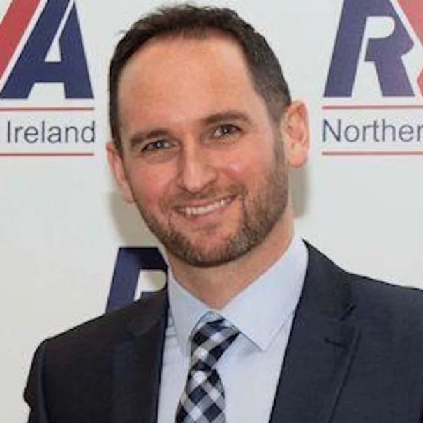 Portrait of a smiling man in business attire, standing in front of a banner with partially visible text.