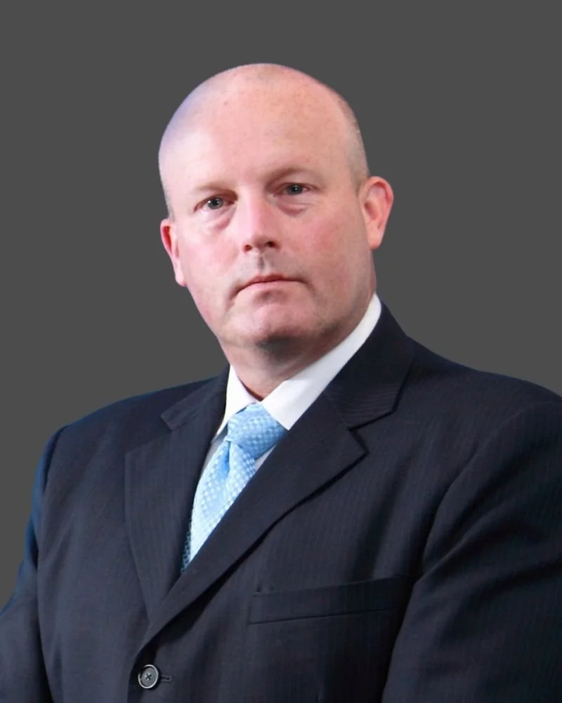 A man in a dark suit, white shirt, and light blue tie against a plain gray background.