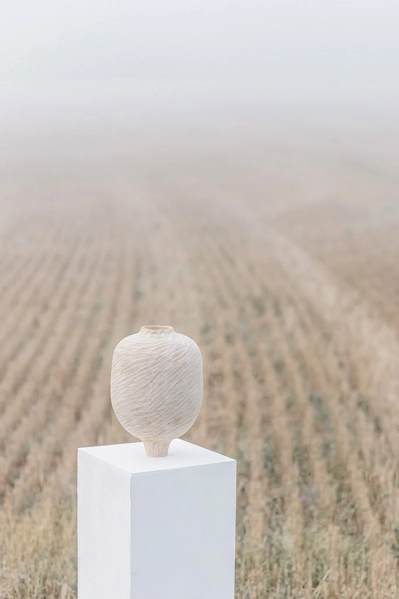 Keraaminen maljakko asetettuna valkoiselle jalustalle pellon keskellä, taustalla sumuinen ja avoin maaseutu.