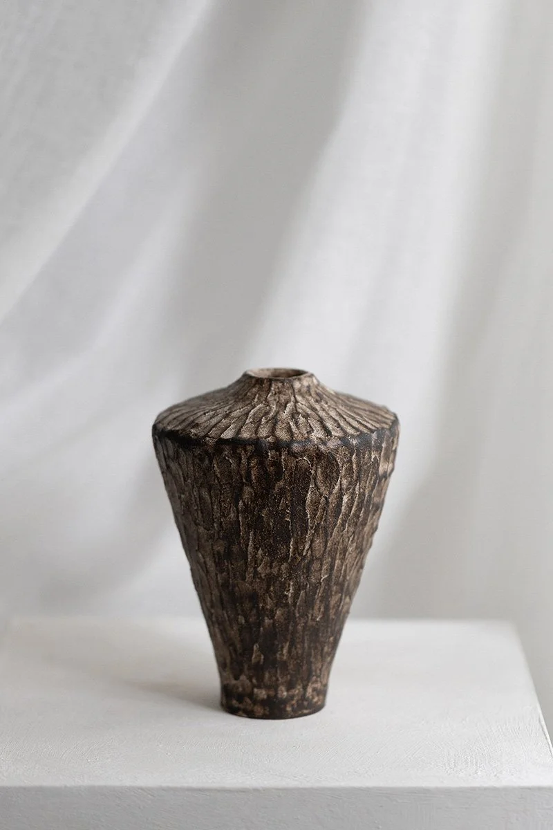 Kuvassa on puisesta saviastiaan muistuttava esine, jonka pinta muistuttaa puun kuoren pintaa, ja se on asetettu valkoiselle alustalle taustalla vaaleat harmaat sävyt.