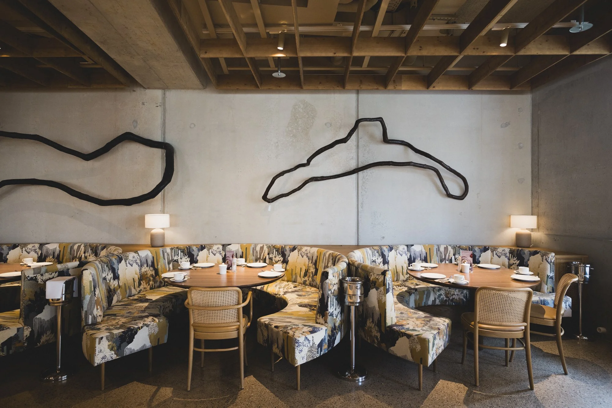 Ein gemütlicher Essbereich mit mehreren Tischen und gepolsterten Stühlen, Wanddekorationen in Form von schwarzen Holzinstallationen auf einer hellen Wand, Lampen auf dem Hintergrund