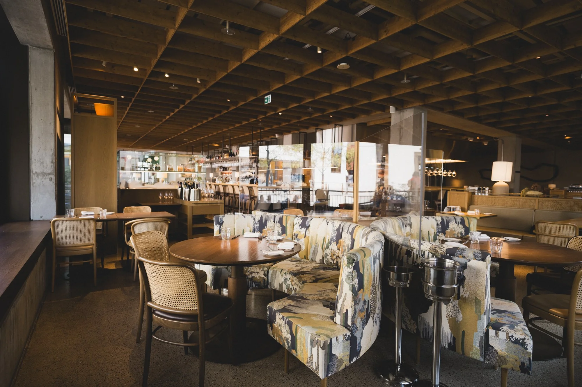 Innenaufnahme eines modernen Restaurants mit bunten Polstern, Holztischen und Bar im Hintergrund, viel natürliches Licht.