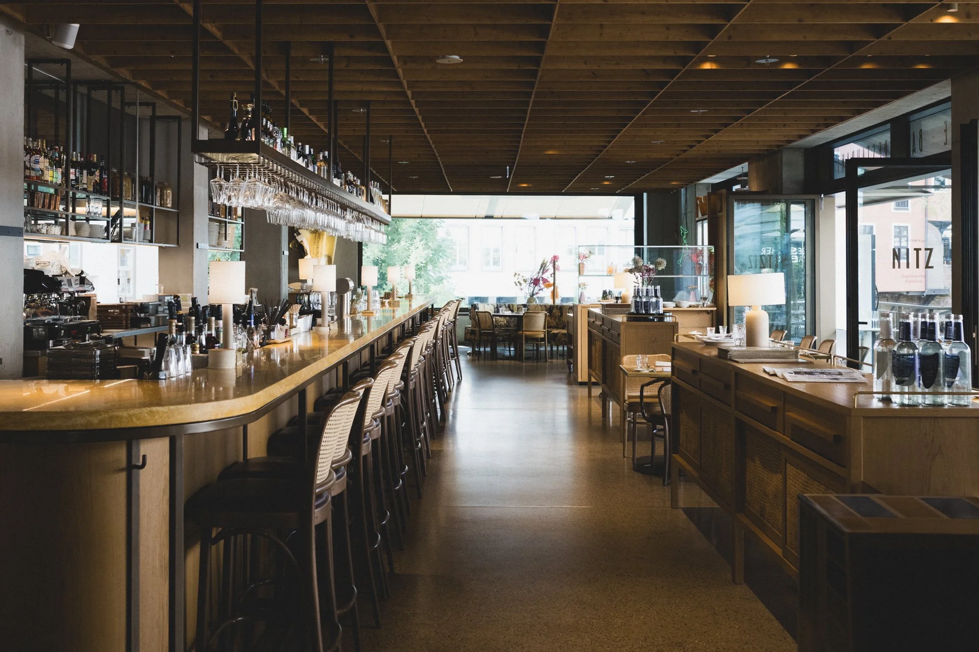 Innenaufnahme eines modernen Restaurants mit Bar, Holzmöbeln, Stühlen, Tischen mit Besteck und Gläsern, Blumen, und großen Fenstern, die Tageslicht hereinlassen.