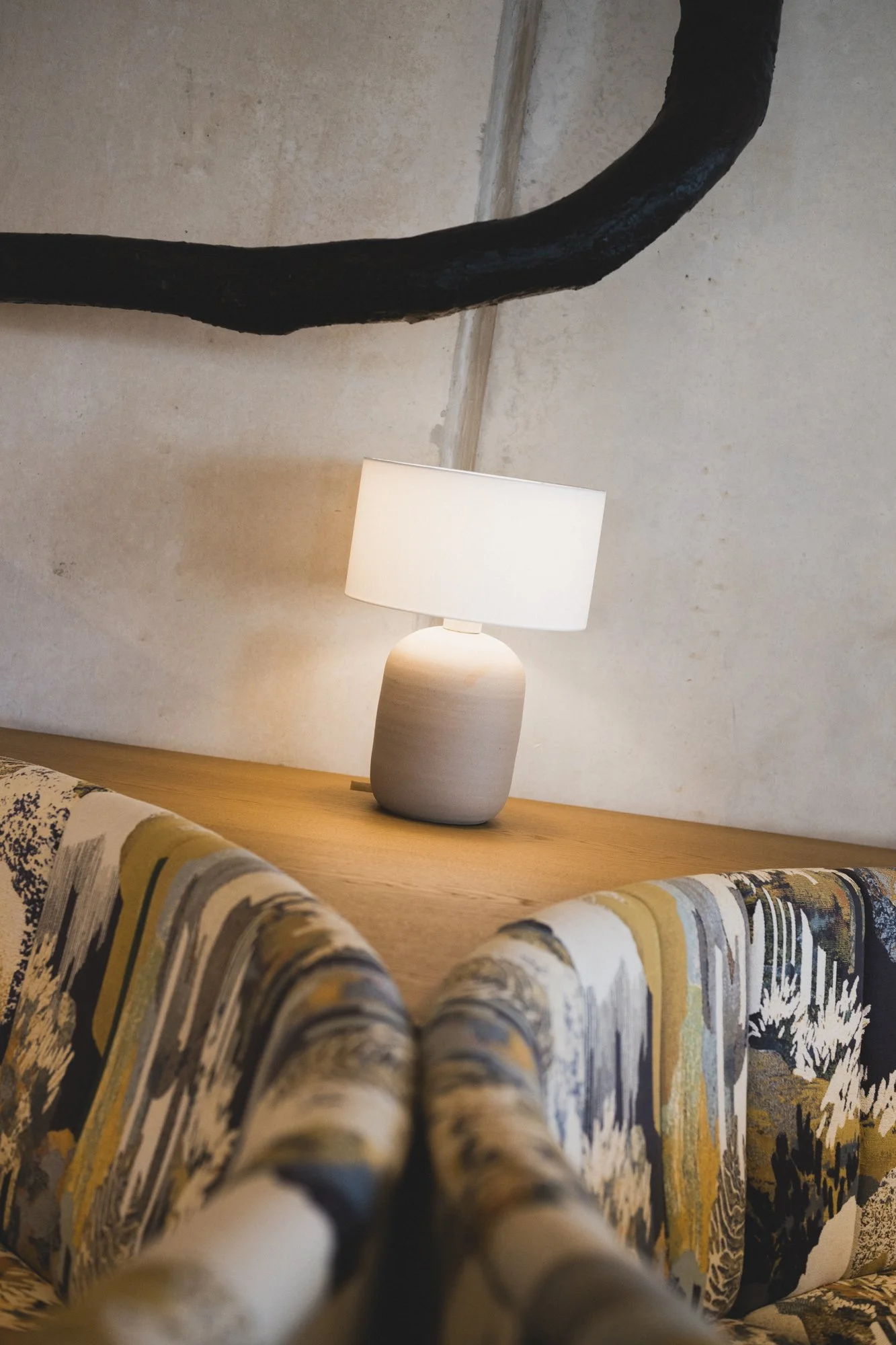 Ein Tisch mit einer weißen Lampe, eine Wand mit einem schwarzen Ast darüber, und eine gepolsterte Couch im Vordergrund.