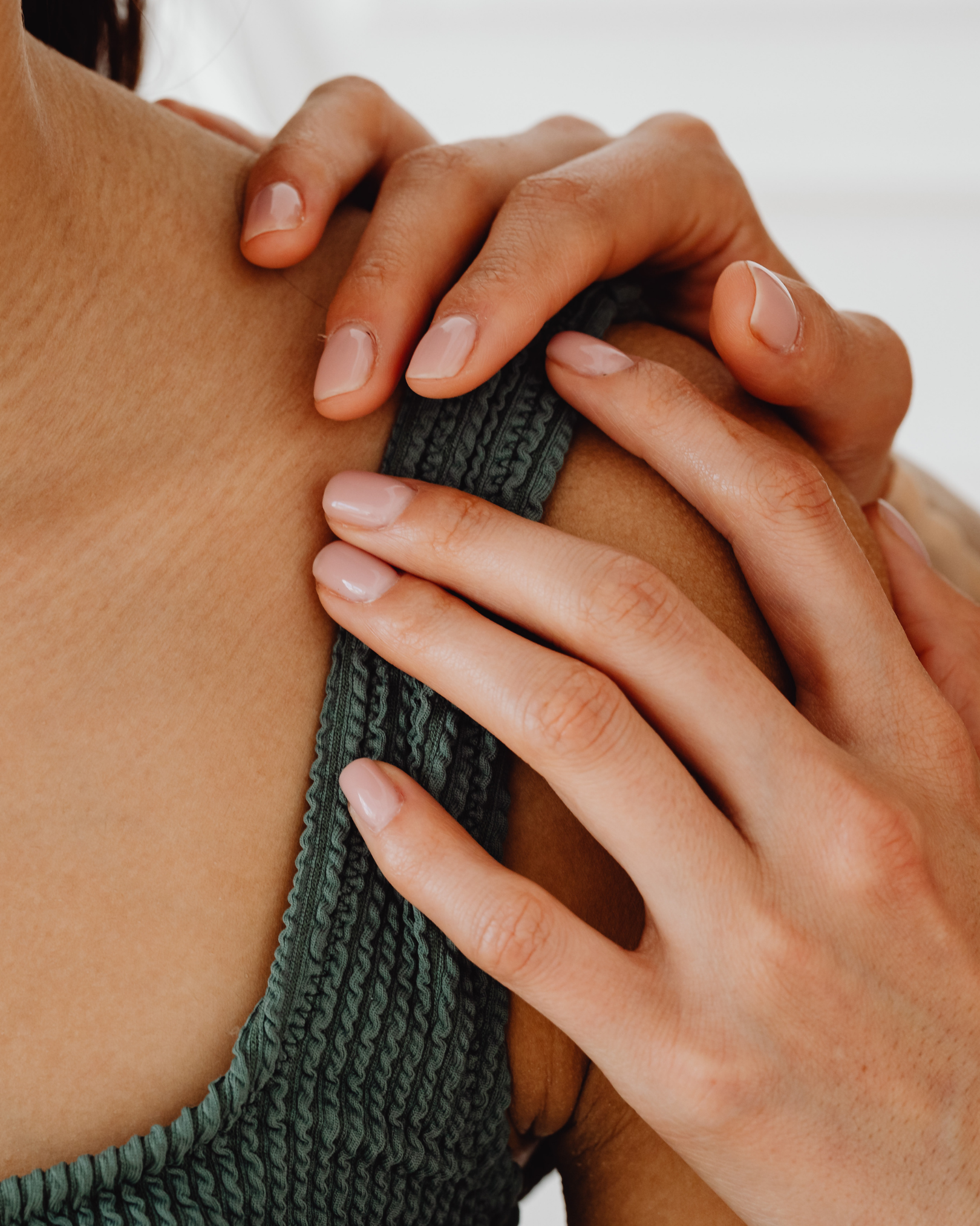 A close-up of a person with tanned skin, wearing a dark green textured tank top with a strappy design, lightly touching their shoulder with another person's hand that has a lighter skin tone and neatly manicured nails.