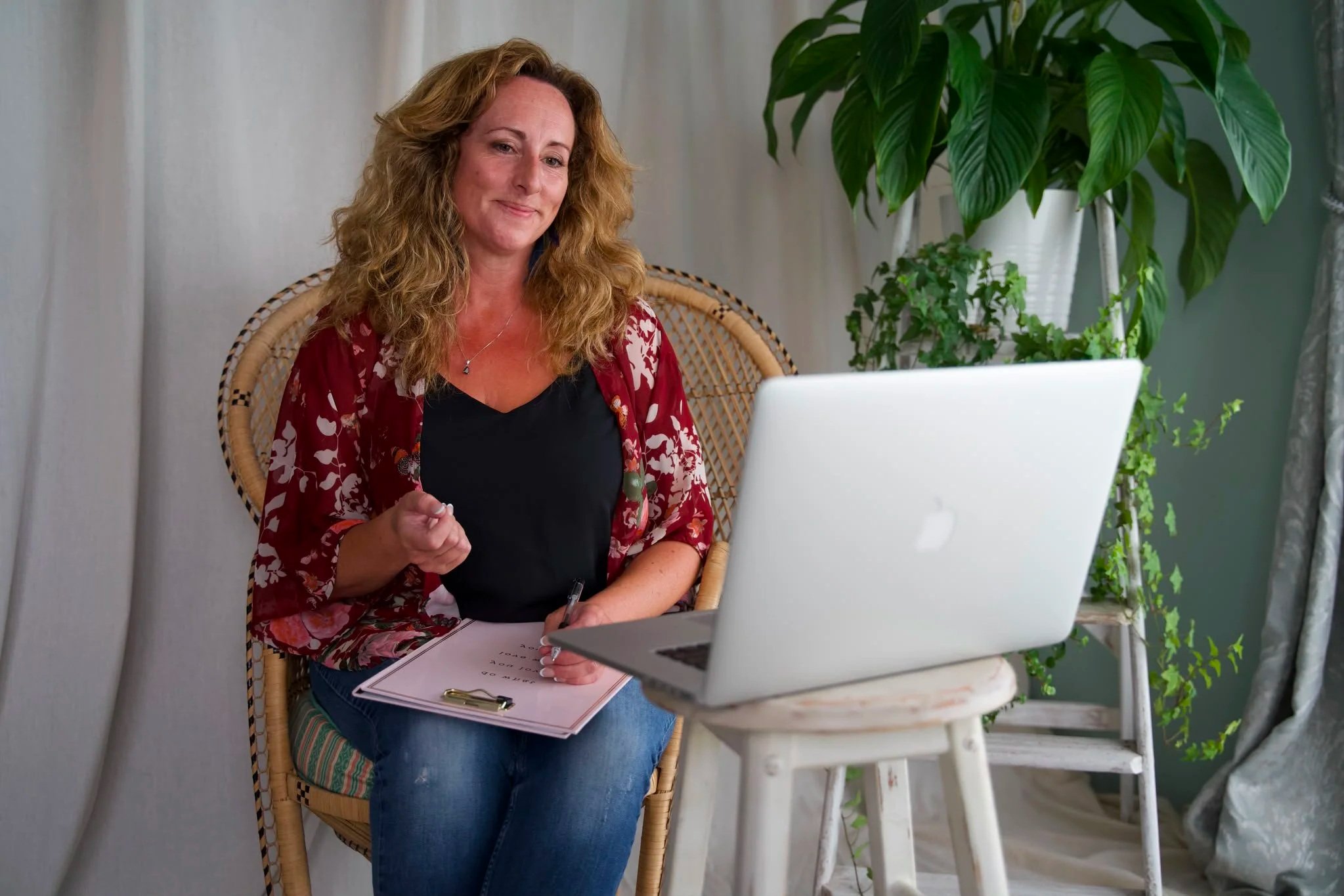 A woman sitting outdoors at a small table with a laptop, holding a pen, with green plants behind her.