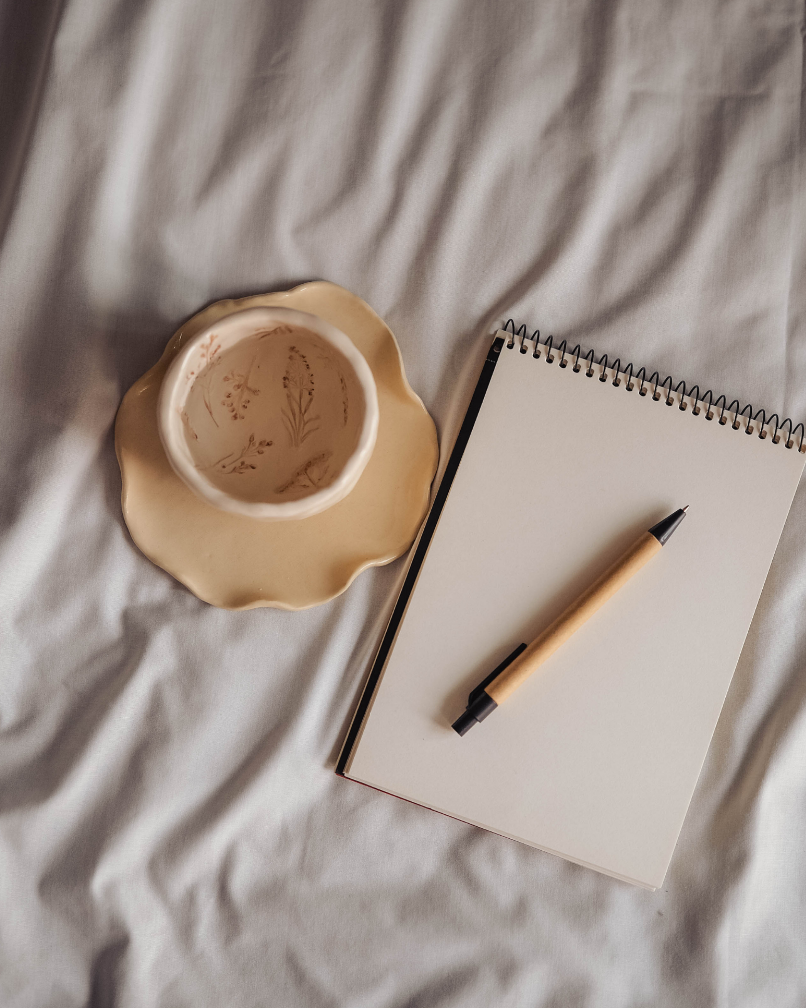An overhead view of a white notepad with black binding, a beige marker, a decorative ceramic cup with a floral pattern on a beige plate, all placed on a white fabric surface.