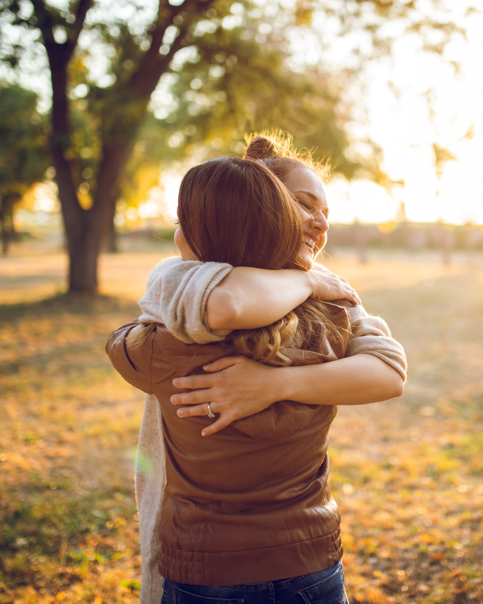 Two women hugging outdoors in a park during sunset, with fall foliage in the background.