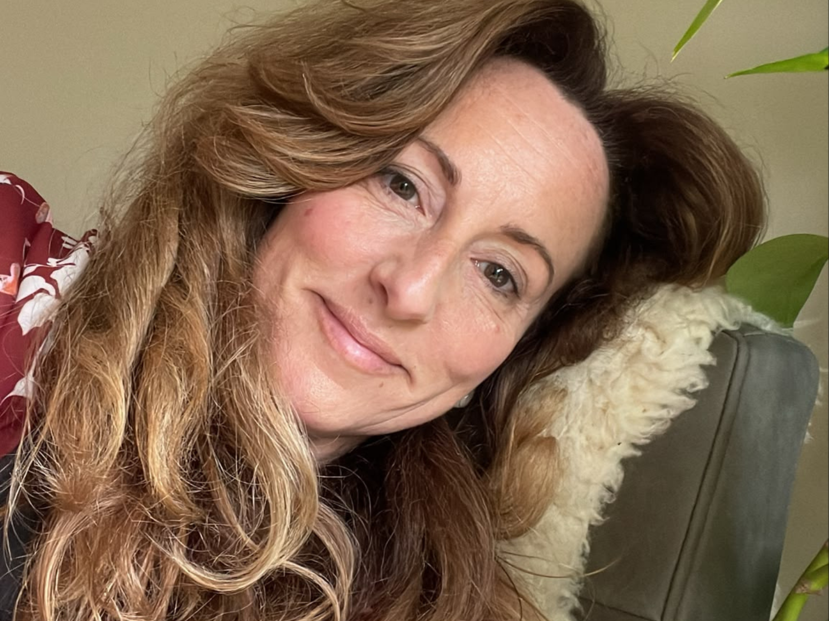 A woman with long, wavy, light brown hair, smiling, seated on a chair with a beige fur blanket. Green plants are visible in the background.