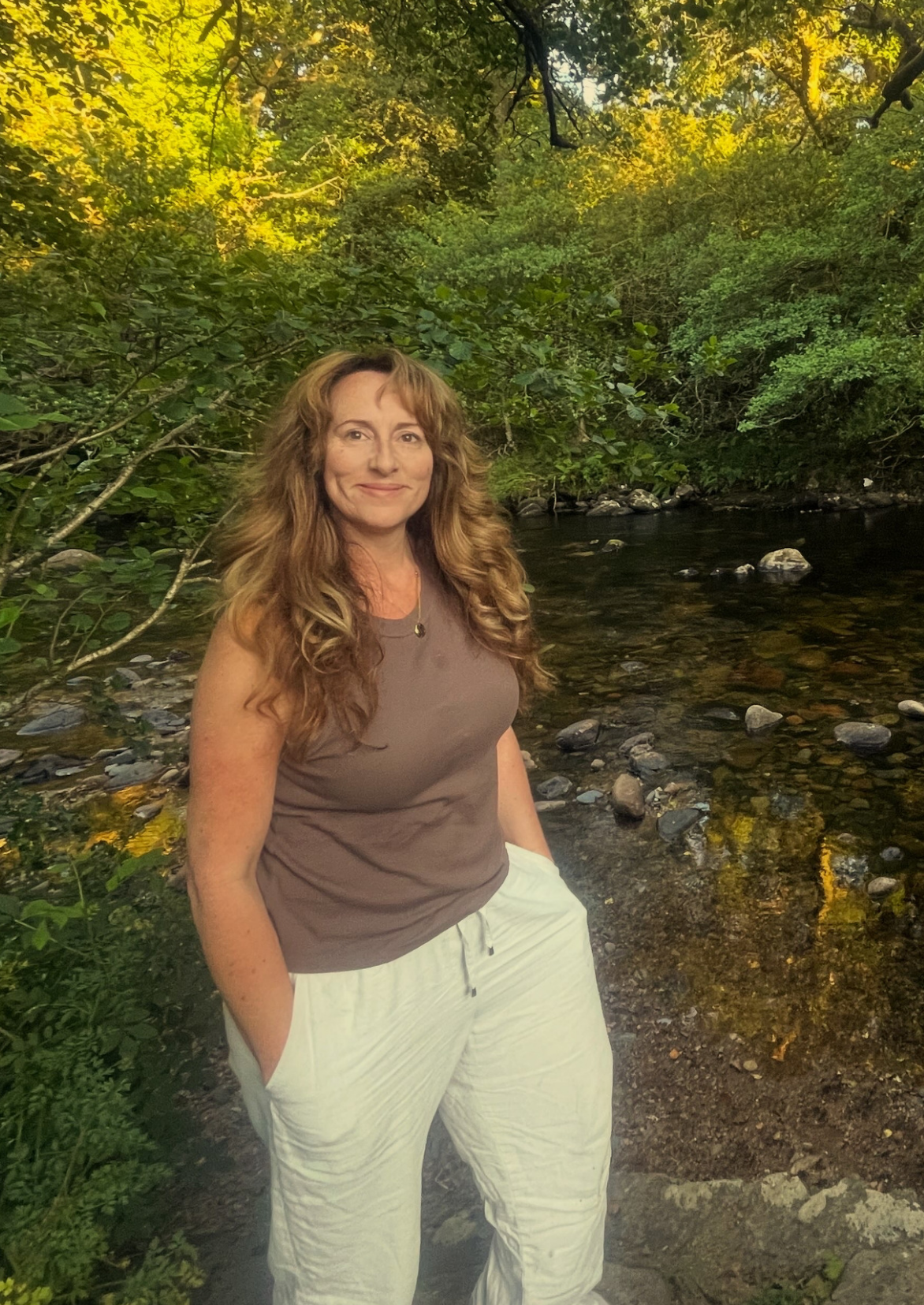 A woman with long, wavy brown hair, wearing a sleeveless brown top and white pants, standing by a stream in a lush, green forest with trees and rocks in the background.