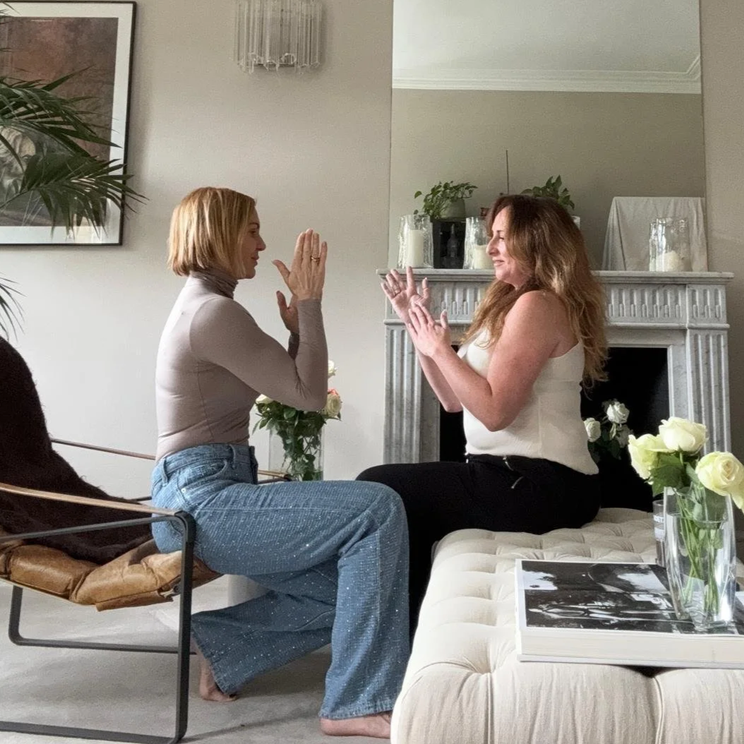 Two women are sitting face-to-face in a living room, engaging in a joyful moment with clapping and smiling at each other. One woman with blonde hair is dressed in a beige turtleneck and jeans, while the other woman has long curly hair and is wearing a white sleeveless top and black pants. The setting includes a beige tufted sofa, a table with white roses, and a fireplace with plants on top.