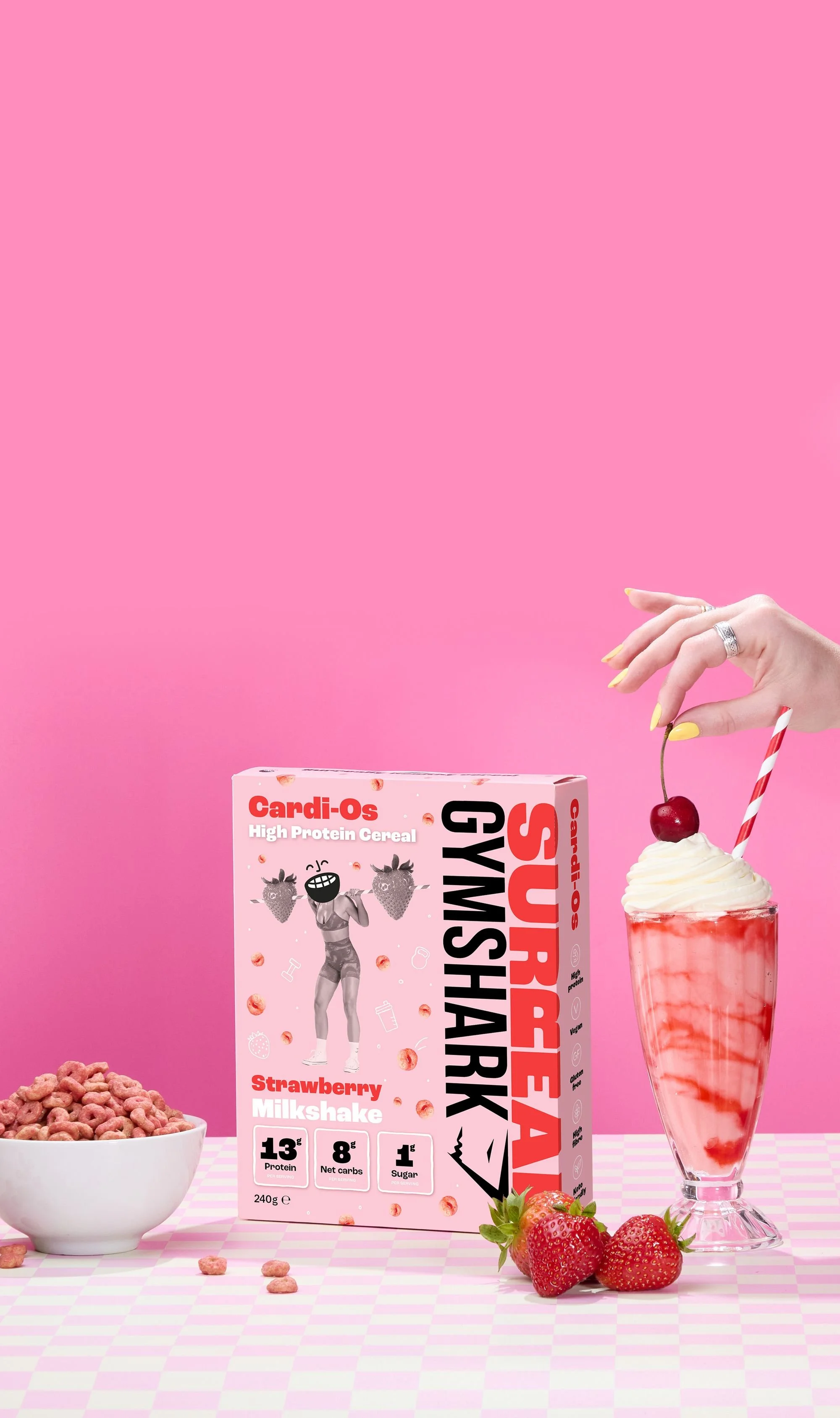 Box of Surreal Gymshark Cardi-Os high protein cereal, strawberry flavor, with a bowl of cereal, strawberries, and a milkshake on a pink checkered surface.