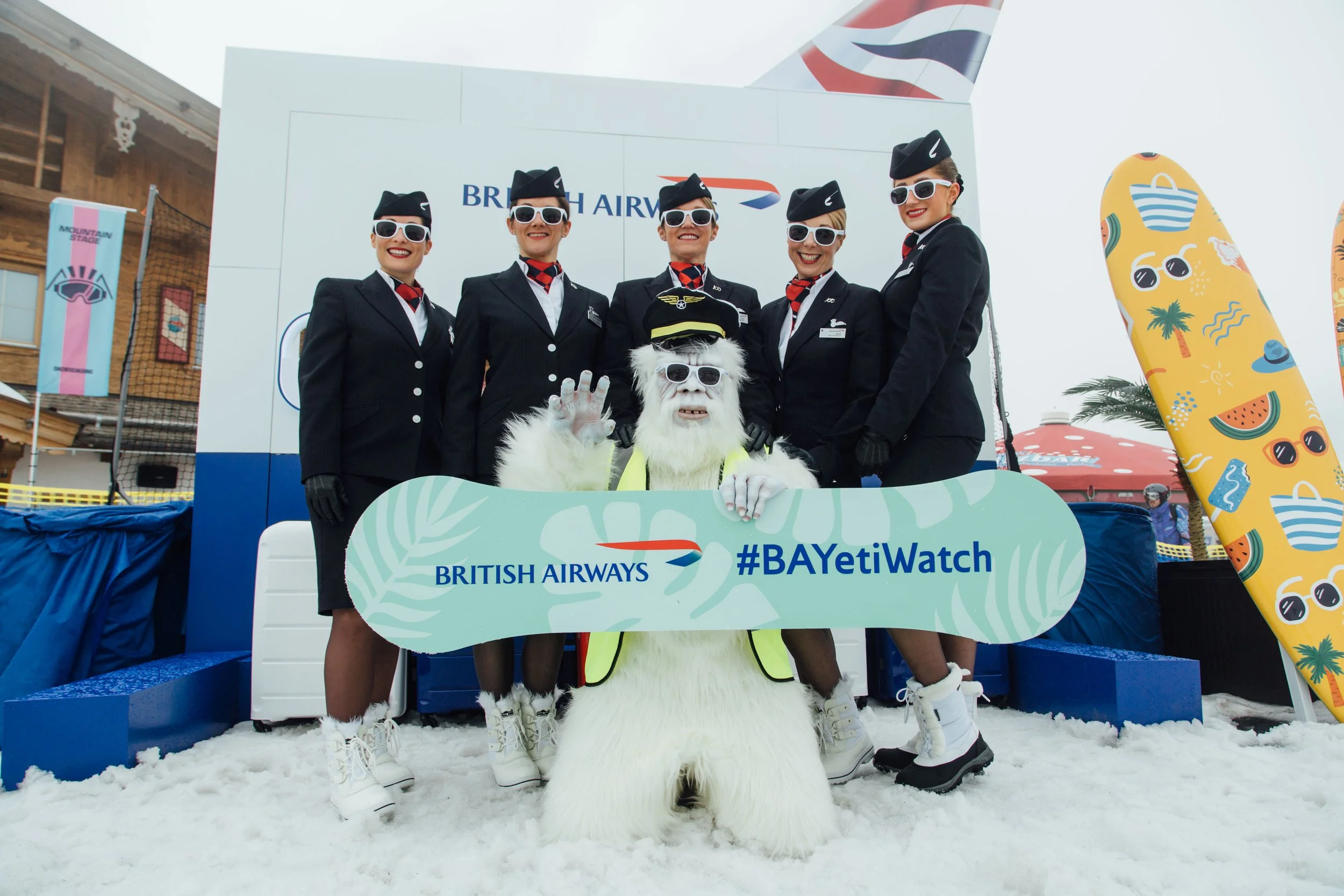 Group of British Airways crew with a person in yeti costume holding a branded snowboard promoting #BAYetiWatch, on a snowy backdrop.