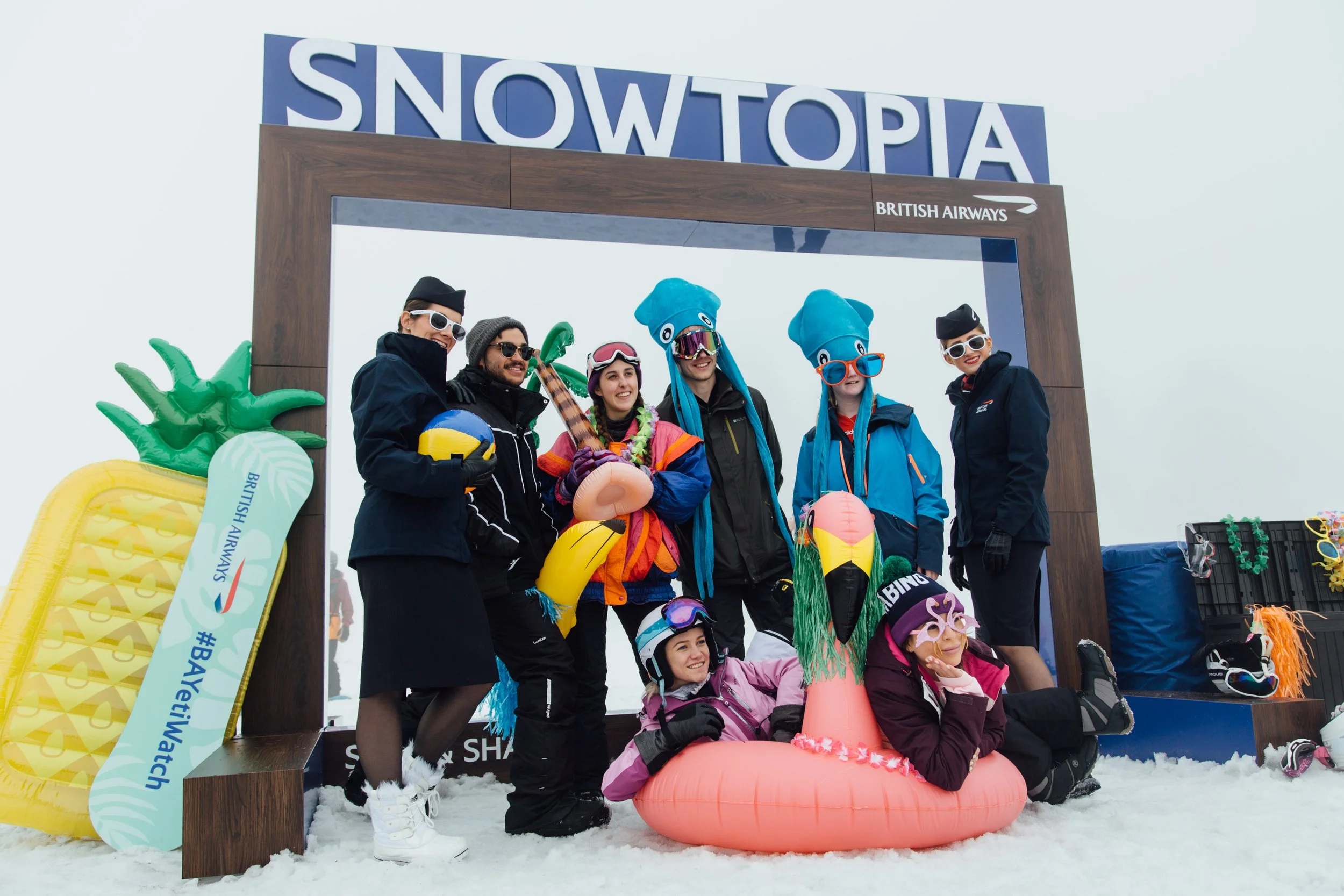 Group of people in winter clothing posing in front of a "Snowtopia" sign, surrounded by inflatable pool toys and wearing funny hats and glasses.