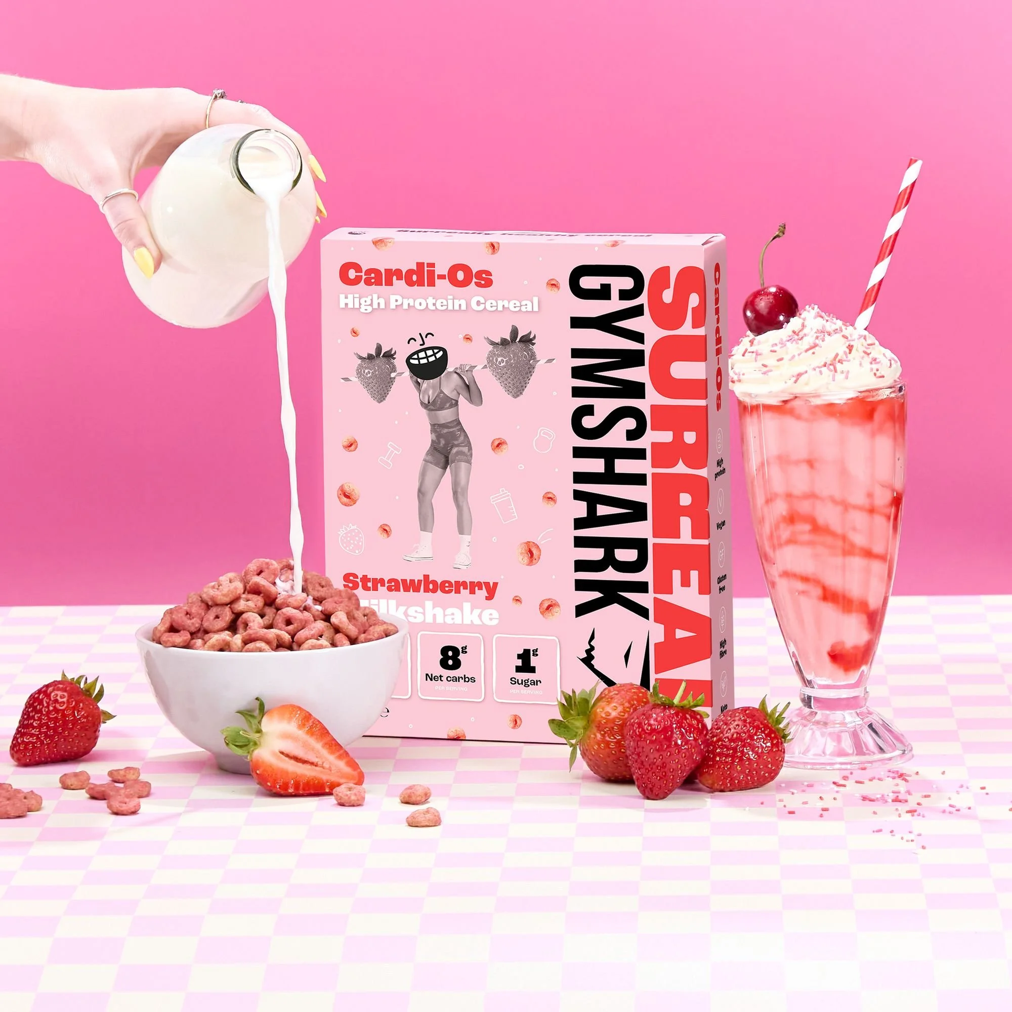Cereal box labeled 'Cardi-Os High Protein Cereal Strawberry Milkshake' with a bowl of pink cereal, milk being poured, fresh strawberries, and a strawberry milkshake on a pink checkered table.
