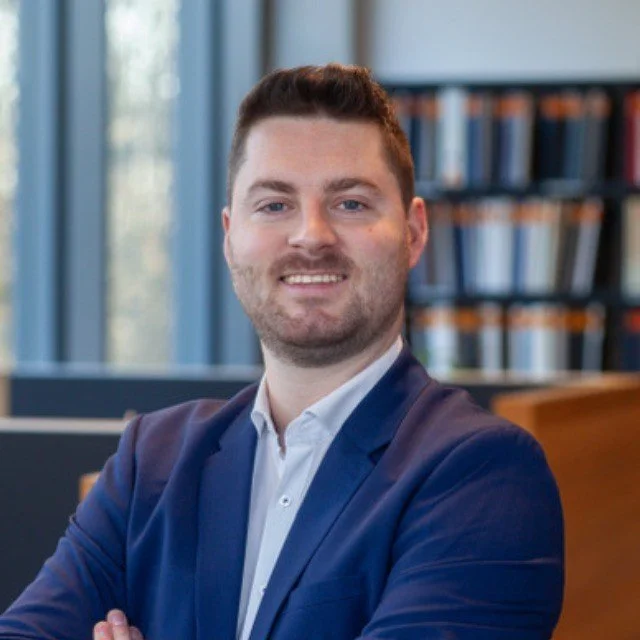 Ein Mann in einem blauen Anzug und weißem Hemd steht in einer Büroumgebung, im Hintergrund Bücherregale.