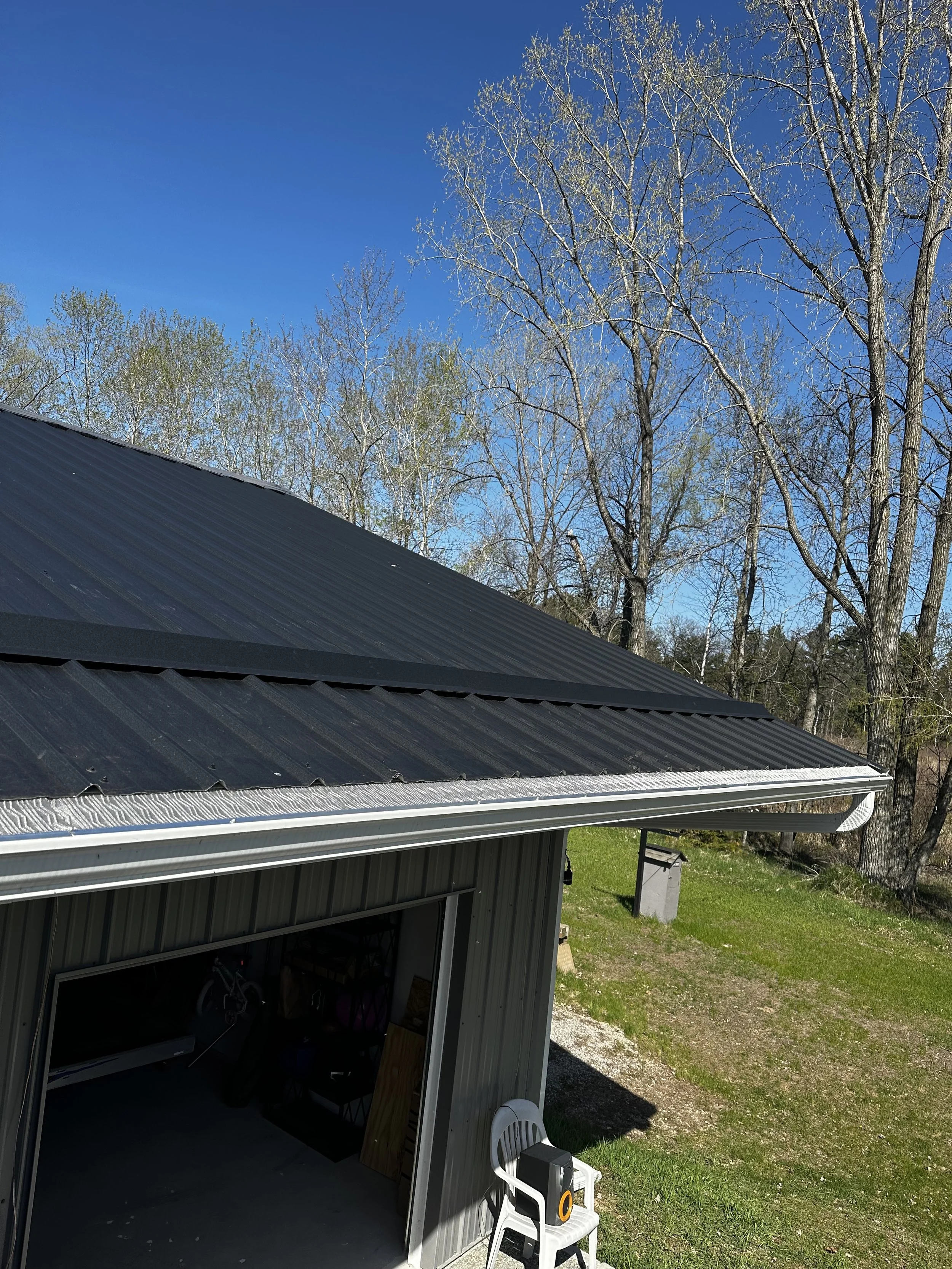 View of a metal building with a black roof and white trim, surrounded by grass and trees in the background, with a partly open garage door, a white chair, and some equipment outside under a clear blue sky.