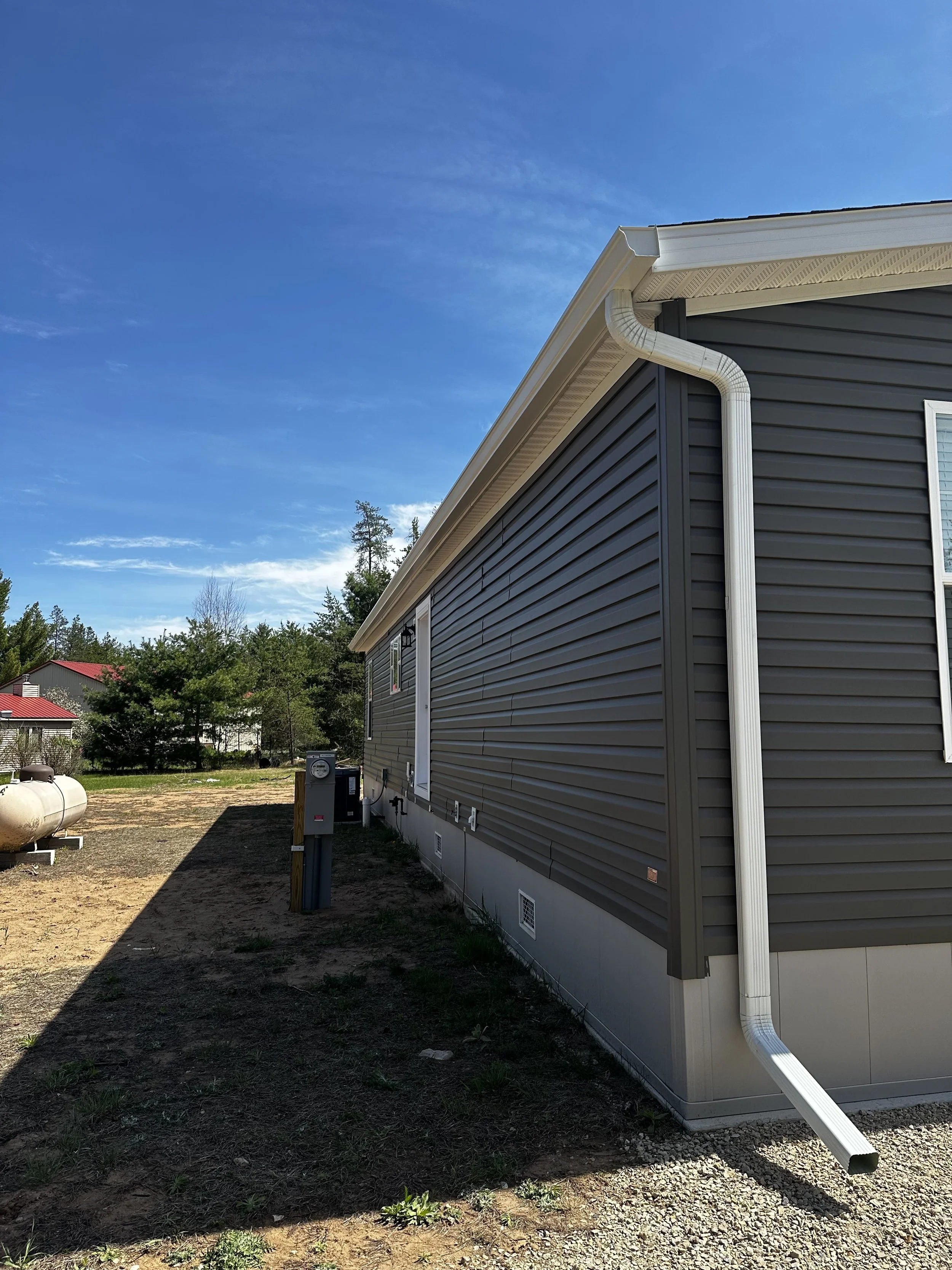 Side of a house with gray vinyl siding, white gutters, and downspouts, under a blue sky with some clouds.