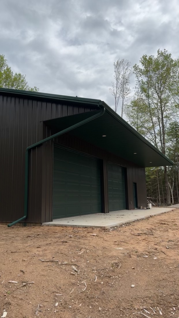 New black metal building with two garage doors and a small door, surrounded by dirt and trees, under a cloudy sky.