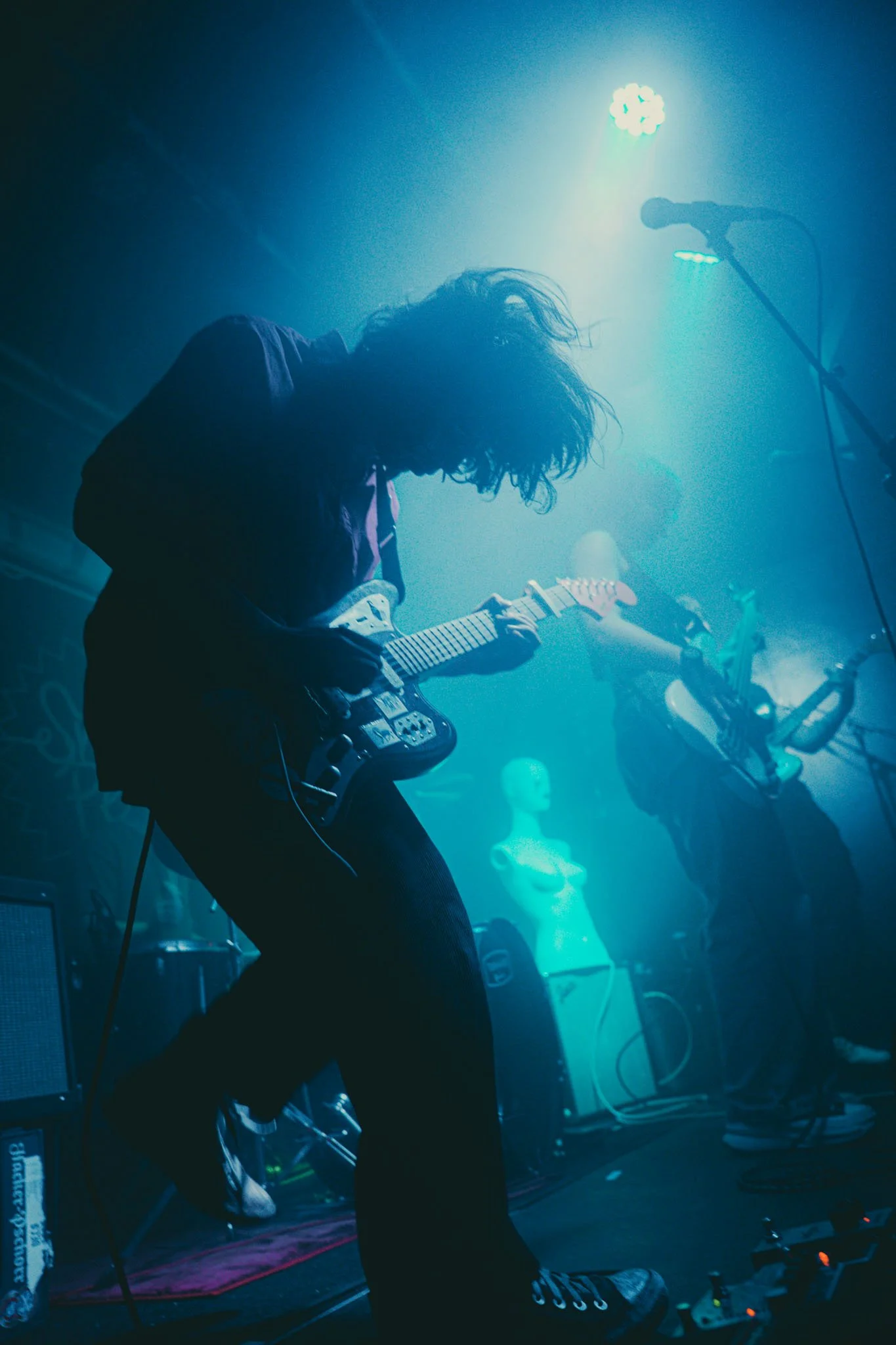 A musician with curly hair playing an electric guitar on stage with blue lighting, with a mannequin and another musician in the background.