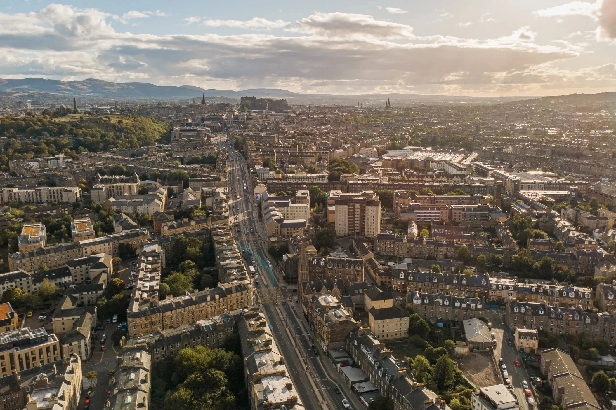 Aerial view of a cityscape with multiple buildings, streets, and green areas, during sunset with partially cloudy sky. Real Estate  Drone Photography Edinburgh