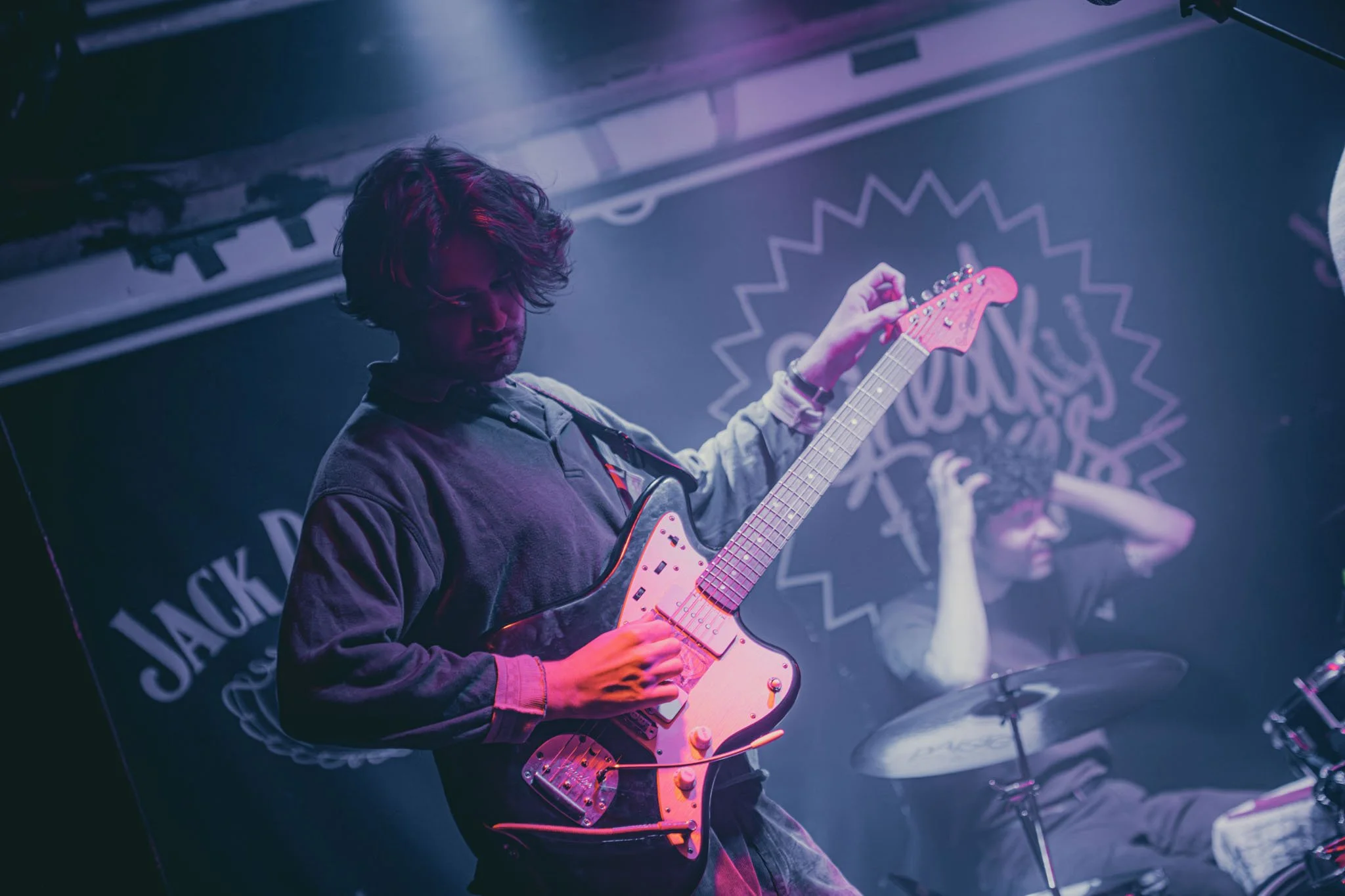 Musician playing electric guitar on stage with drummer in background at Jack Daniel's venue.